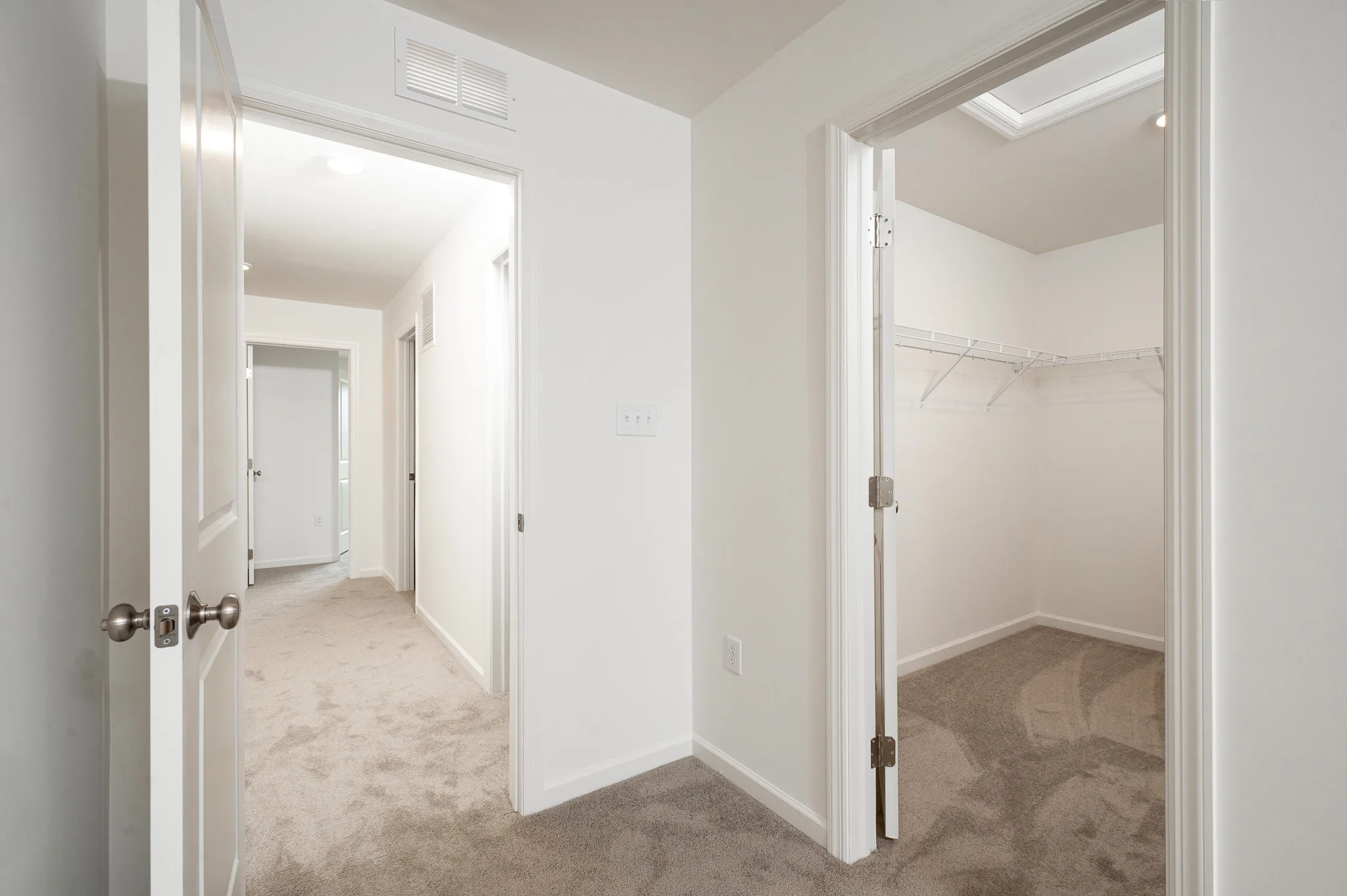 A view of a beige-carpeted hallway with white walls and doors. One open door leads to a walk-in closet with wire shelving, while another door opens to the hallway with additional rooms visible in the distance.