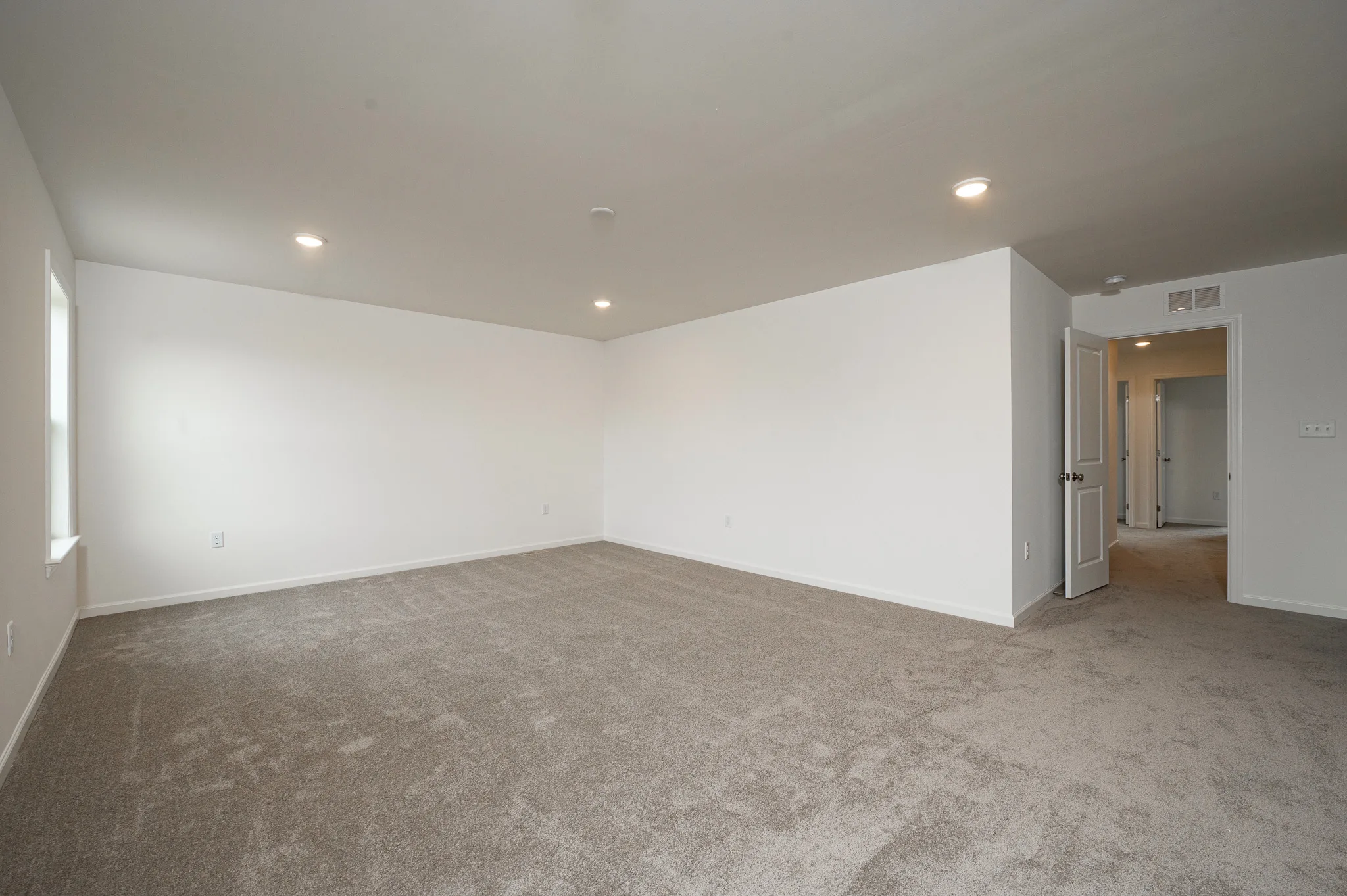 Spacious, empty room with beige carpet, white walls, recessed ceiling lights, a window on the left, and an open doorway leading to a hallway and additional rooms.