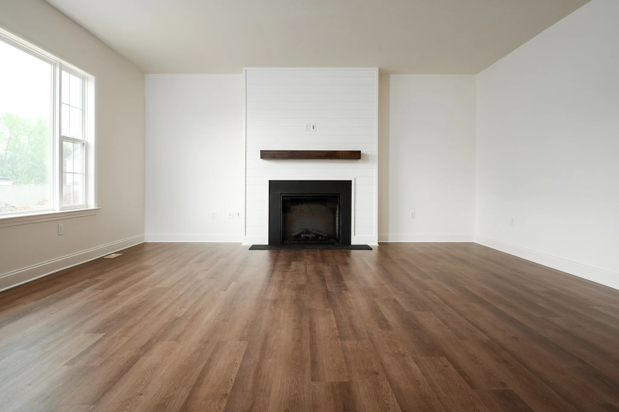 A bright, empty living room with light beige walls, wide windows, wood-style flooring, and a central fireplace with a white shiplap surround and a dark wooden mantel.