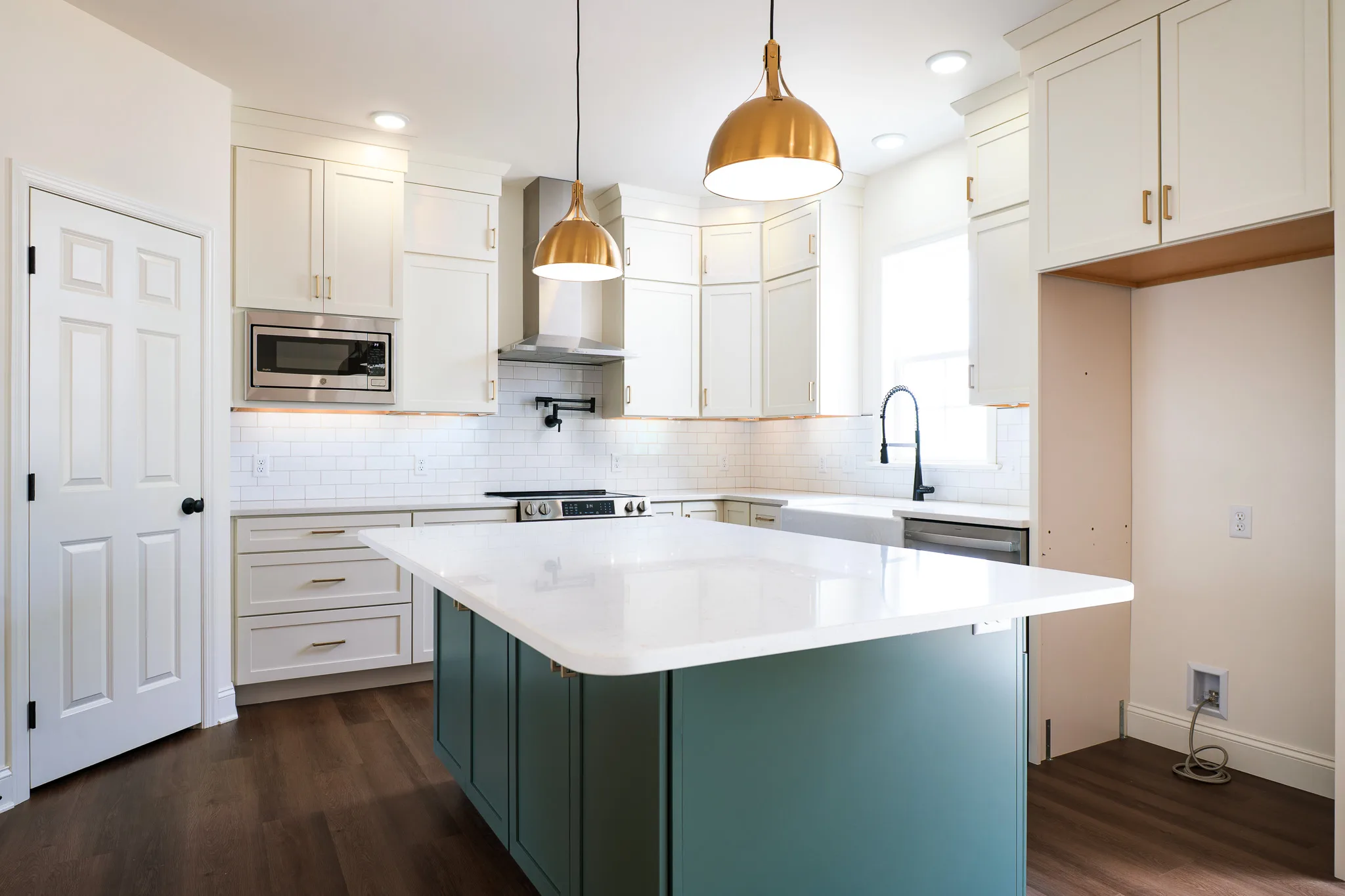 Modern kitchen with white cabinets, gold handles, a teal island with a white countertop, pendant lights, stainless steel appliances, and dark wood flooring. An empty space exists for a fridge next to the island.