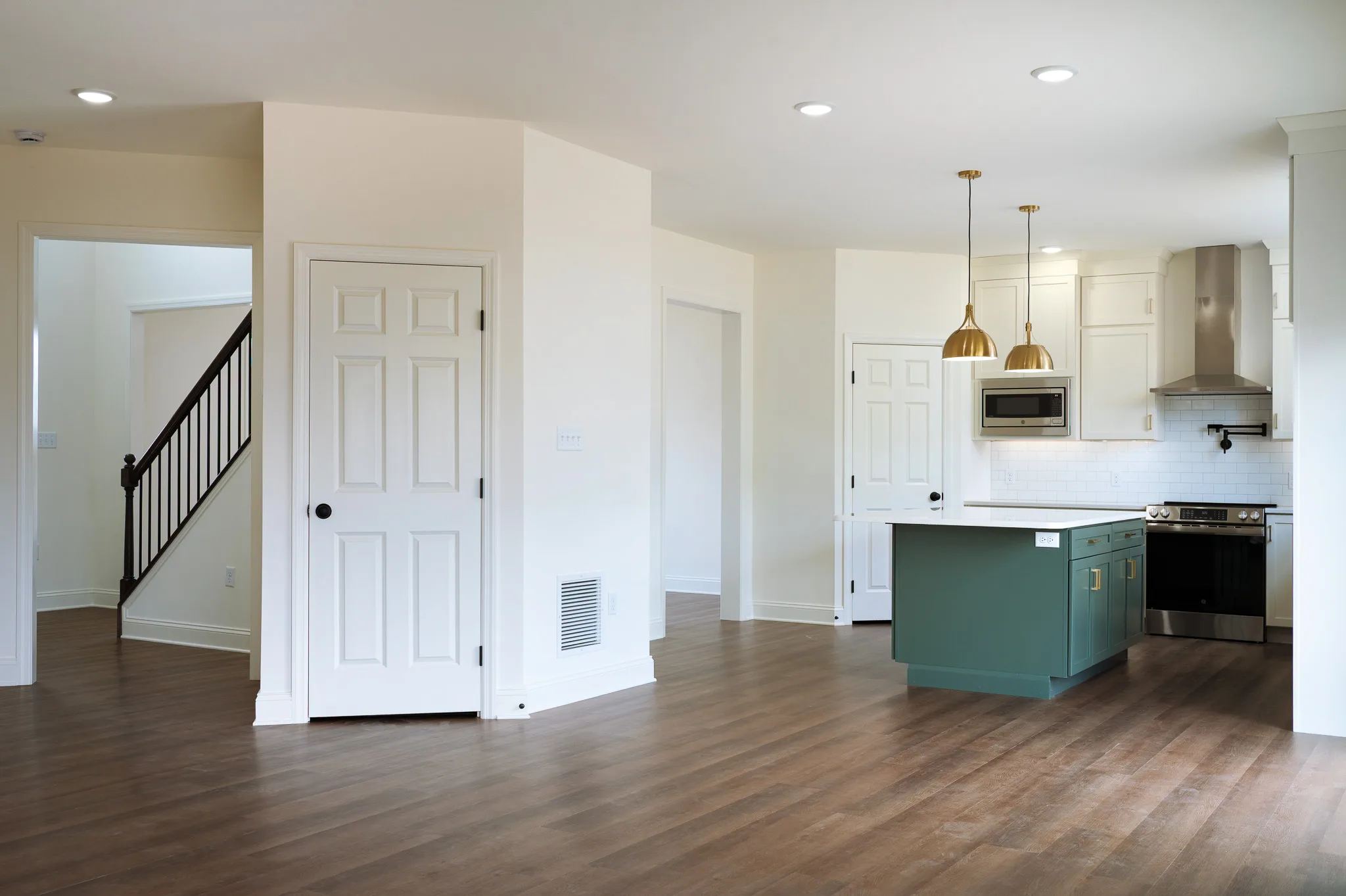 A modern, unfurnished kitchen and living area with wood flooring, white walls, green kitchen island, gold pendant lights, stainless steel appliances, and a staircase with a dark railing in the background.