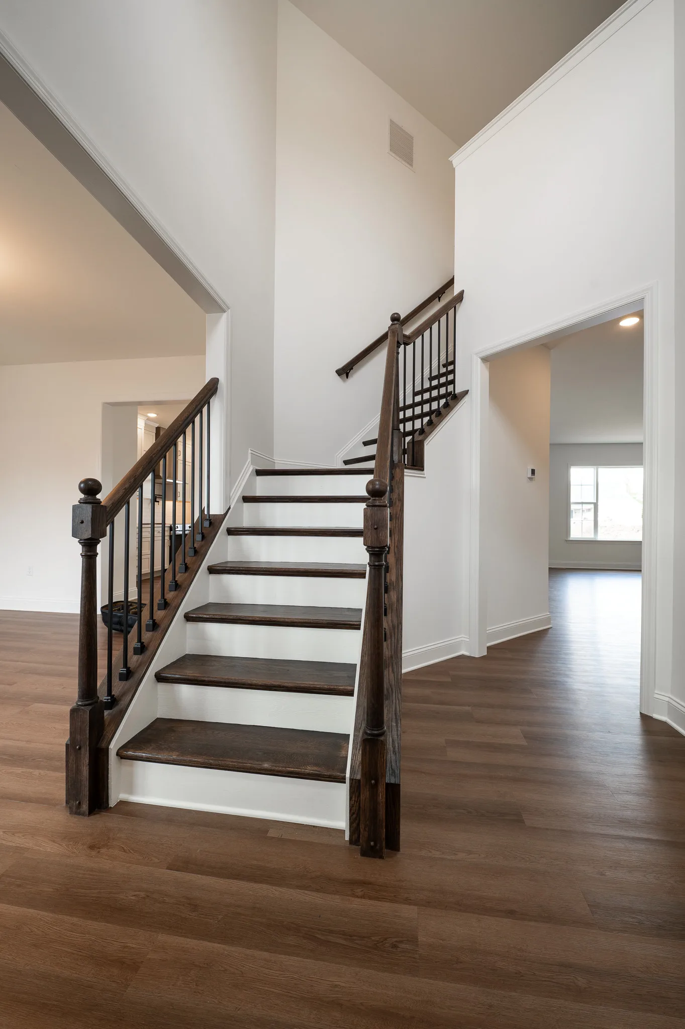 A modern, bright interior features a wooden staircase with dark railings, white walls, and wood flooring. Open doorways reveal additional rooms with large windows and natural light.