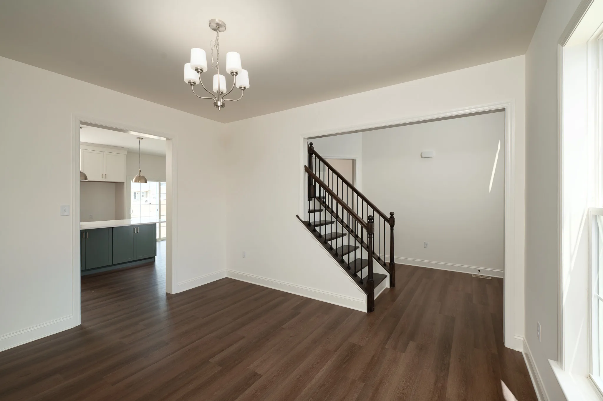 A bright, empty room with light walls, dark wood flooring, a modern chandelier, a staircase with dark railing, and an open doorway leading to a kitchen with teal cabinets and pendant lights.