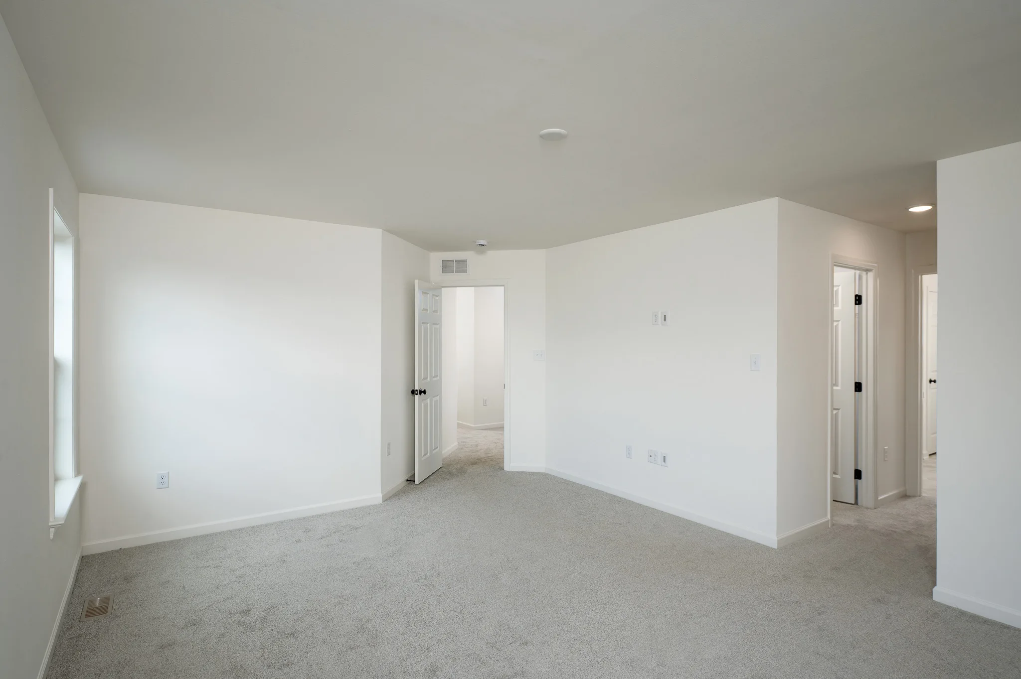 A bright, empty room with white walls, light gray carpet, a window, and multiple doorways leading to adjacent rooms and a hallway. The space is clean and unfurnished.