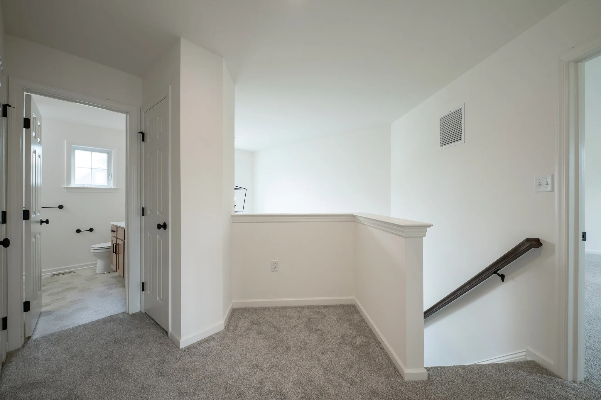 A bright, carpeted upstairs hallway with white walls, a stair railing, doors leading to a bathroom and bedrooms, and natural light entering through multiple windows.