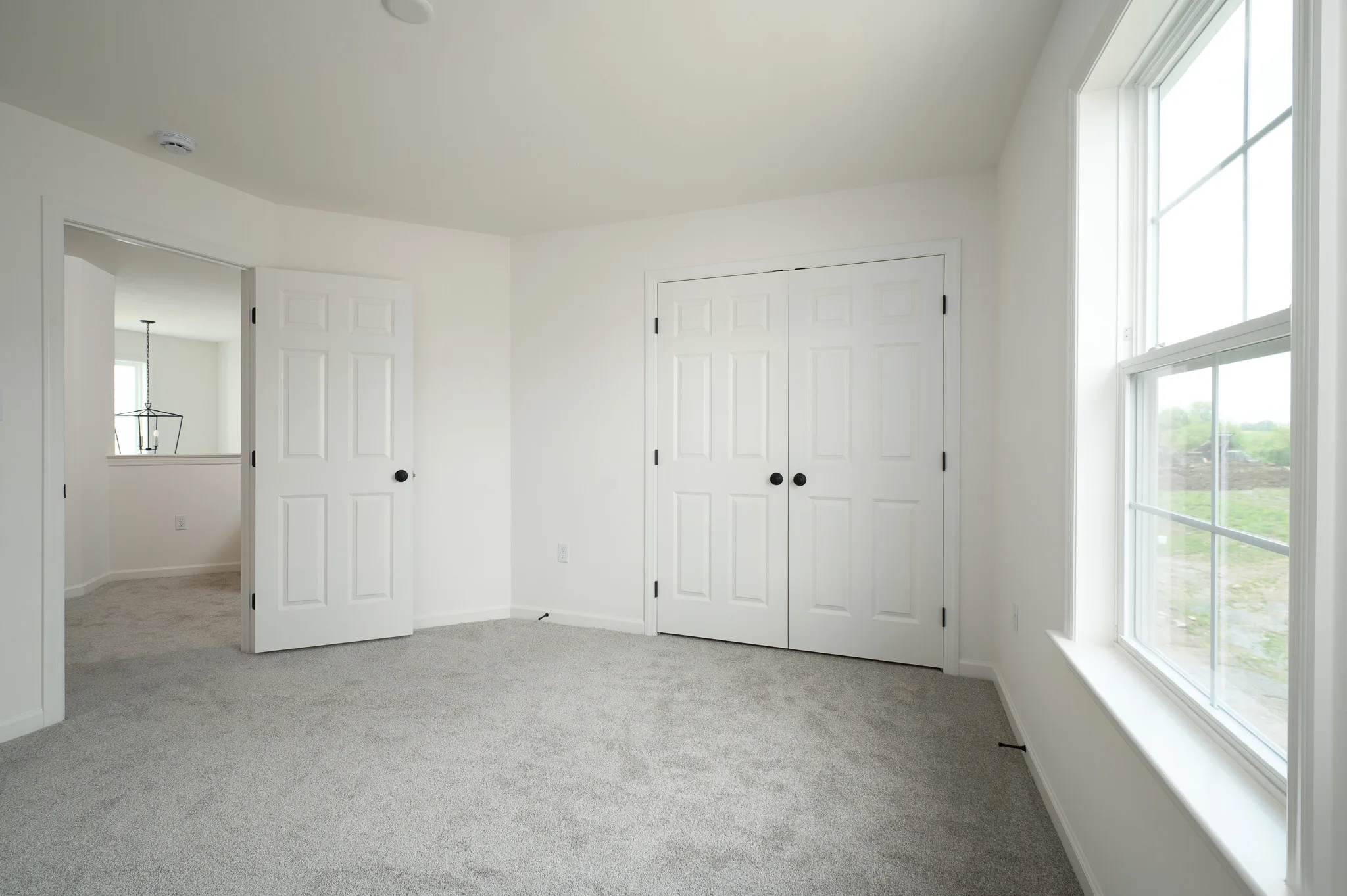 A bright, empty room with light gray carpet, white walls, a large window, double closet doors, and an open door leading to a hallway. Natural light fills the space.
