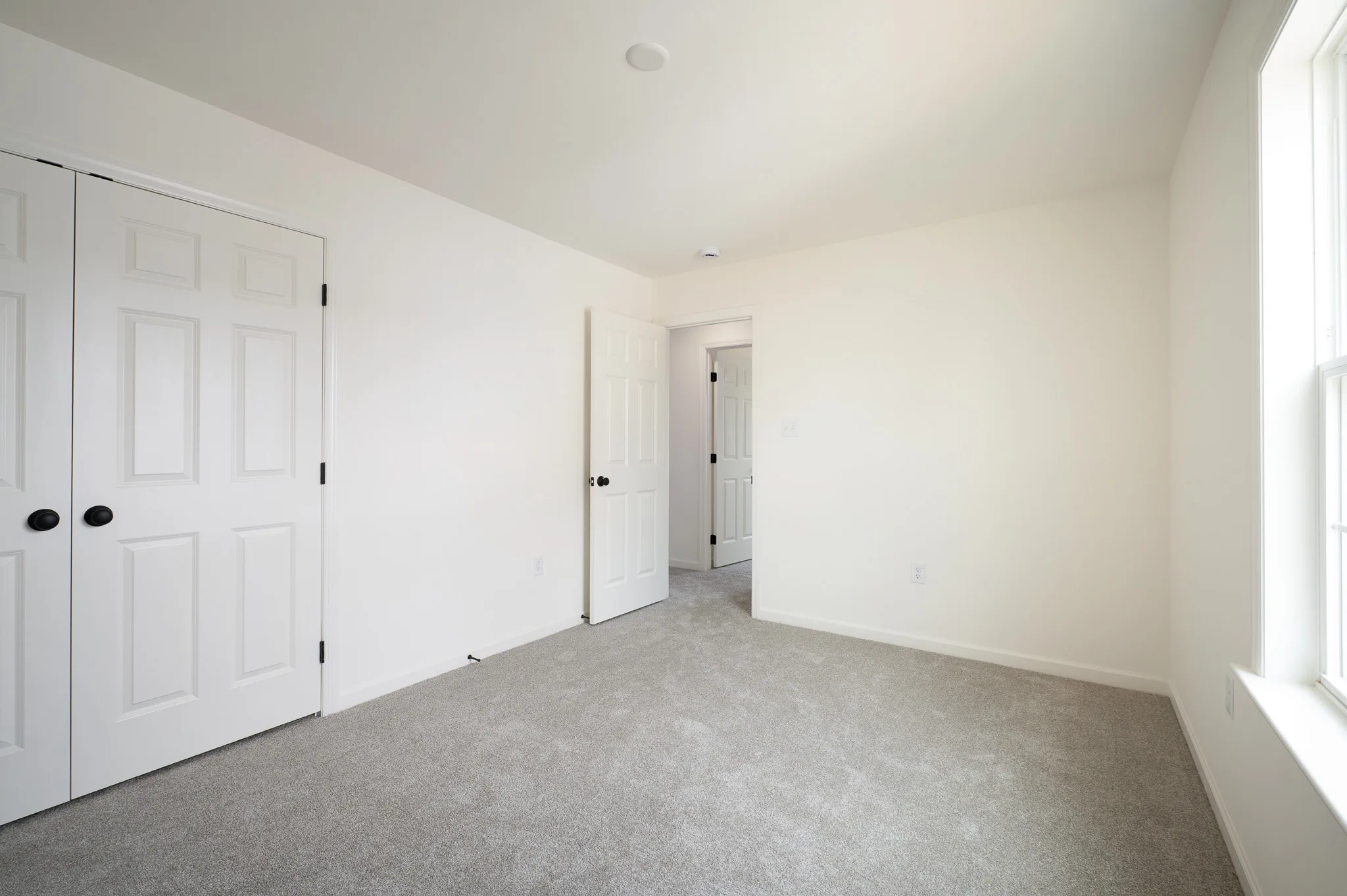Empty bedroom with light gray carpet, white walls, a double-door closet on the left, an open white door leading to a hallway, and a window on the right letting in natural light.