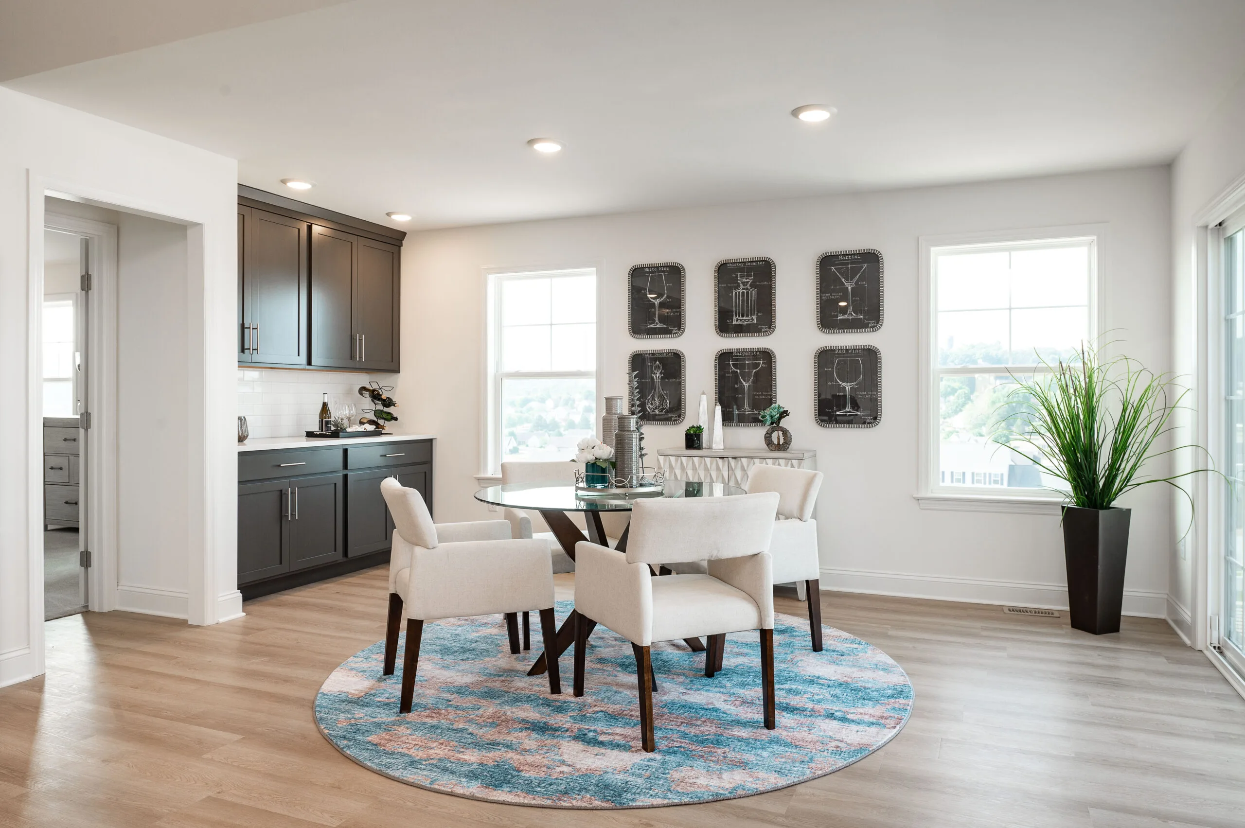 A modern dining area with a round glass table, four white chairs, a blue patterned rug, and gray cabinets. Large windows brighten the space, and wall art and a potted plant add decorative touches.