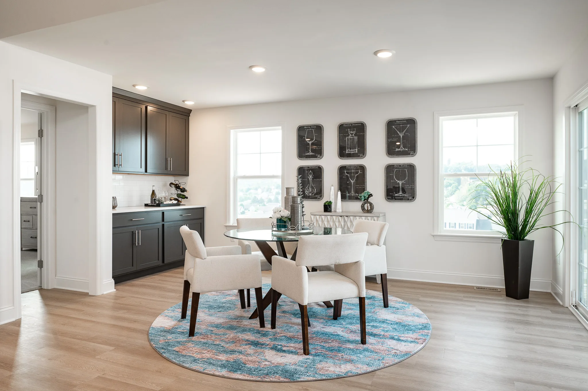 Modern dining area with a round glass table, four white chairs on a blue patterned rug, dark cabinets, and wall art. Large windows let in natural light, offering an Eagle's View of Summergrove. A tall plant sits in the corner by the windows.