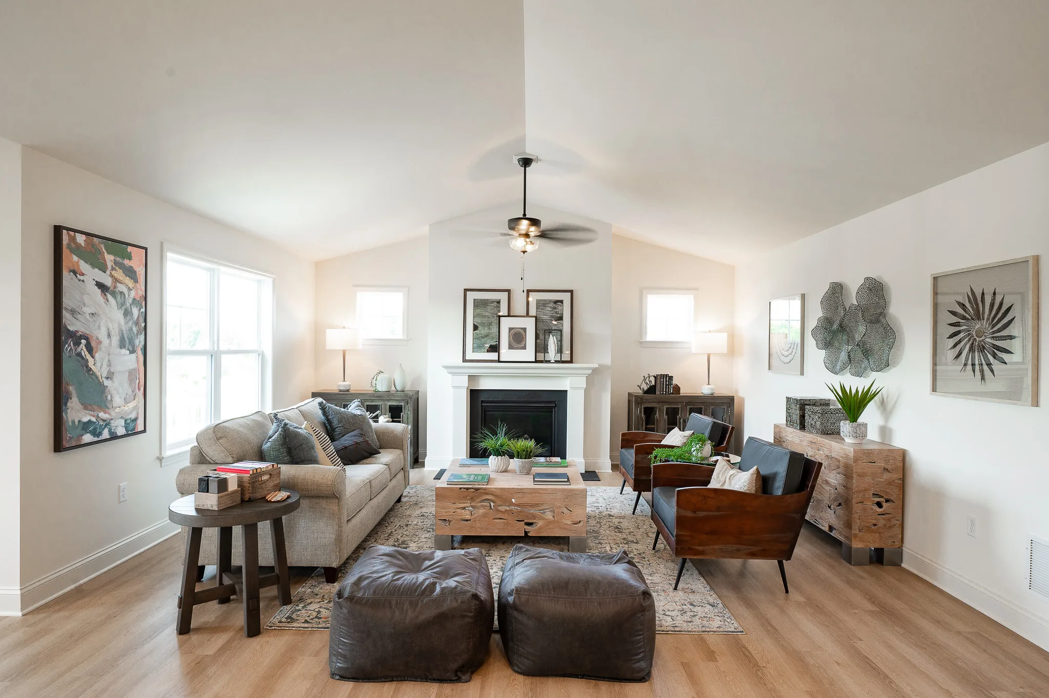 A modern living room at Eagle's View features neutral tones, a gray sofa, two leather chairs, a wooden coffee table, two brown poufs, a fireplace, and contemporary American decor including plants, artwork, and lamps on either side.