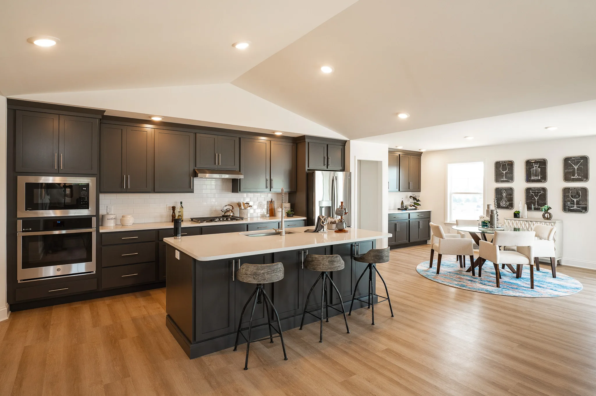 Modern American kitchen with dark cabinetry, a large island with three barstools, and built-in appliances, opening to a bright Eagle's View dining area featuring a round table, white chairs, decorative wall art, and light wood floors throughout.