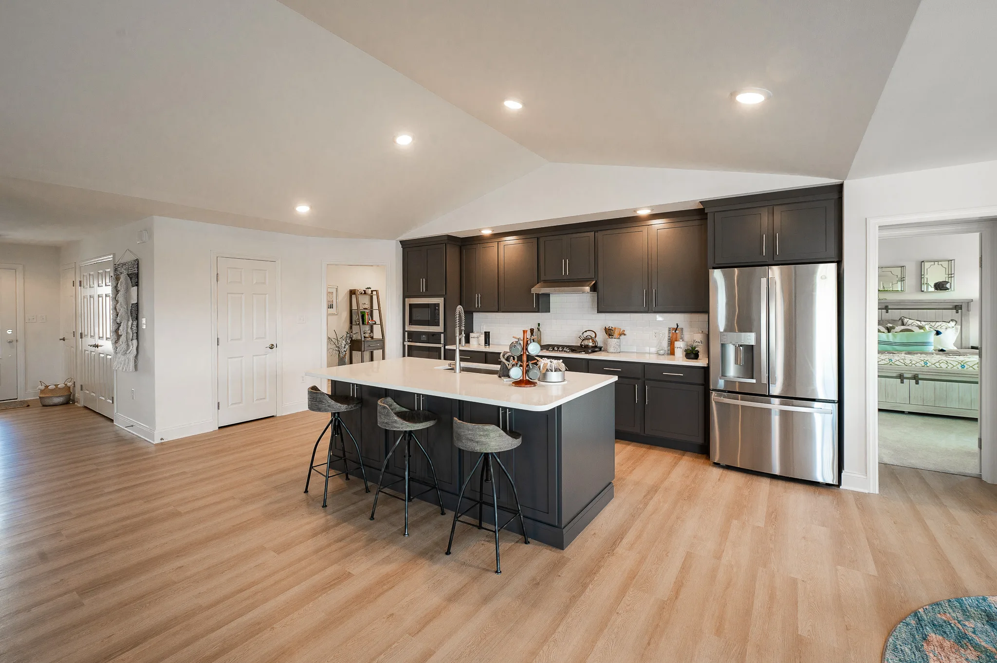 Modern kitchen with dark gray cabinets, stainless steel appliances, and white island with three barstools sits atop light wood flooring. This inviting Summergrove home features an open layout leading to a bedroom and other rooms in the background.