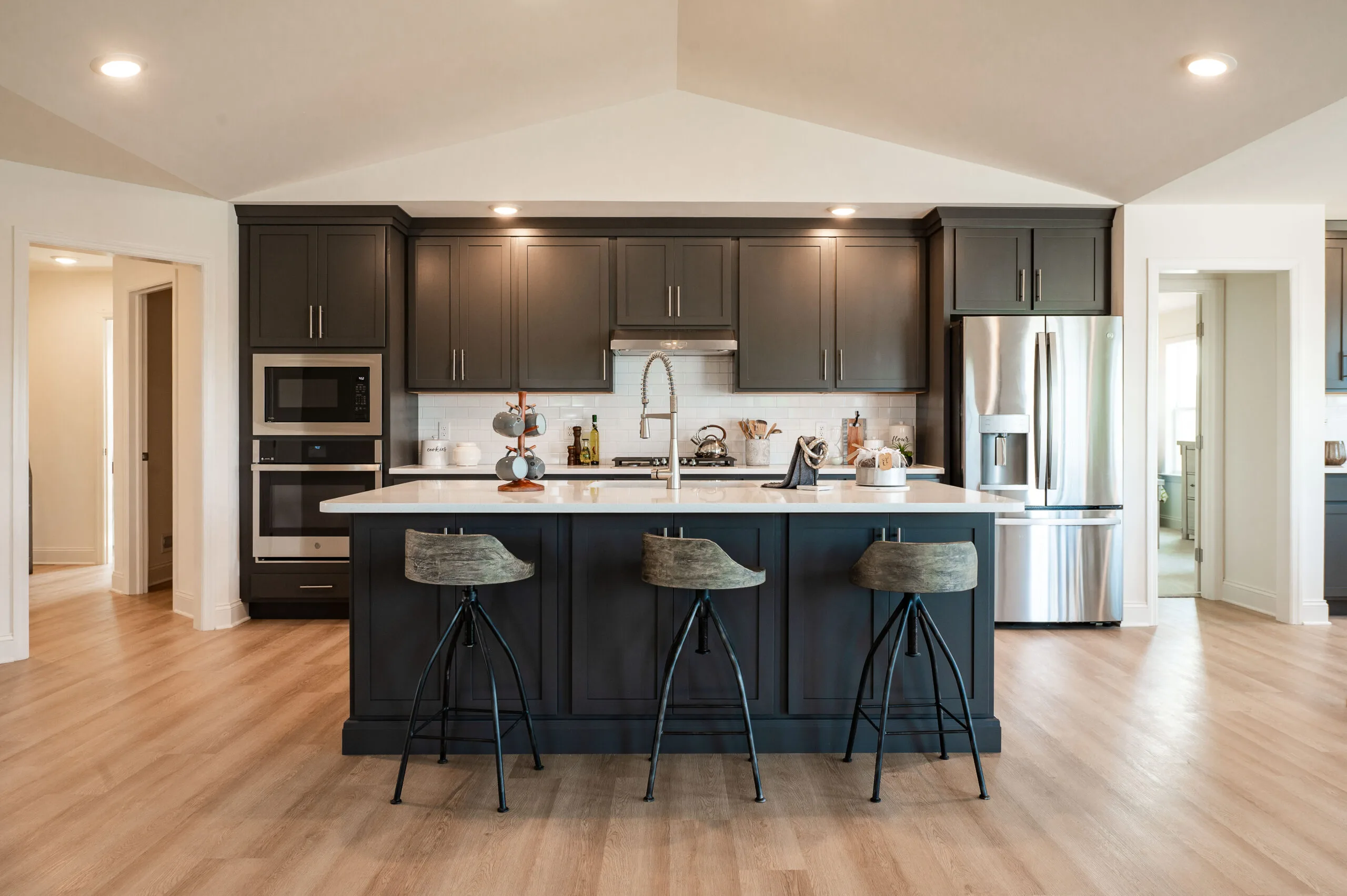 Modern kitchen with dark cabinets, a large center island with three barstools, stainless steel appliances, and light wood flooring. Recessed lighting brightens the open, spacious layout.