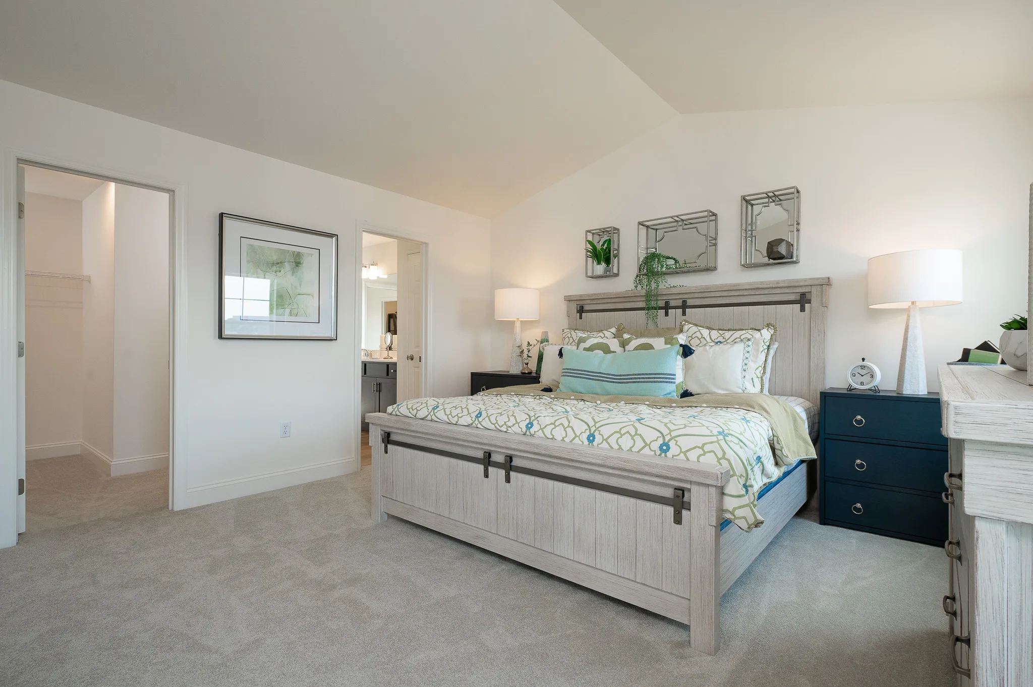 A modern American bedroom in Summergrove with a large bed, decorative pillows, and blue nightstands. Lamps, wall mirrors, and plants adorn the headboard. The white-walled, carpeted room connects to a closet and bathroom.