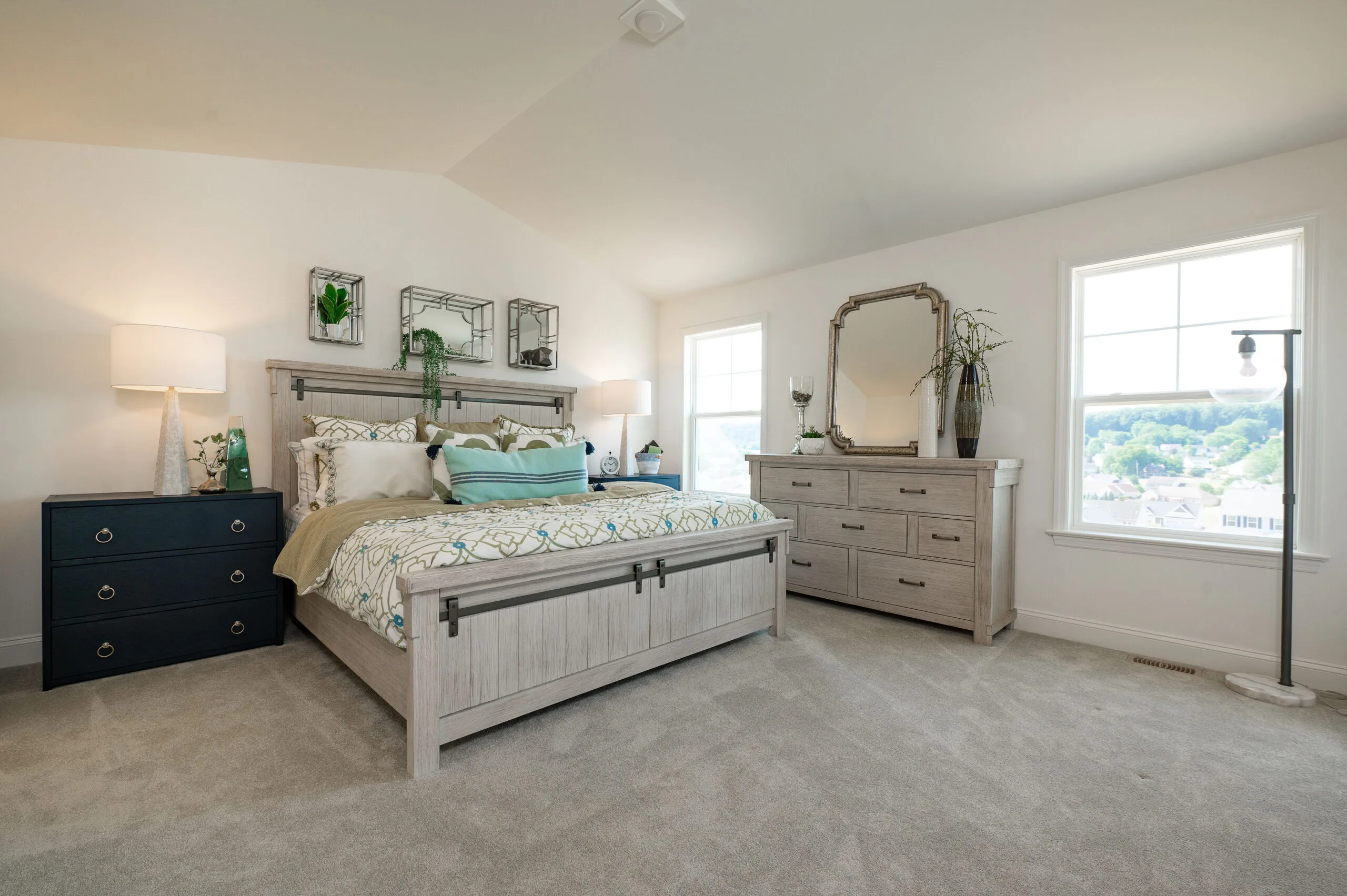 A bright, modern bedroom with a large bed, light wood furniture, blue accents, decorative pillows, two nightstands with lamps, a large dresser with a mirror, and two windows letting in natural light.