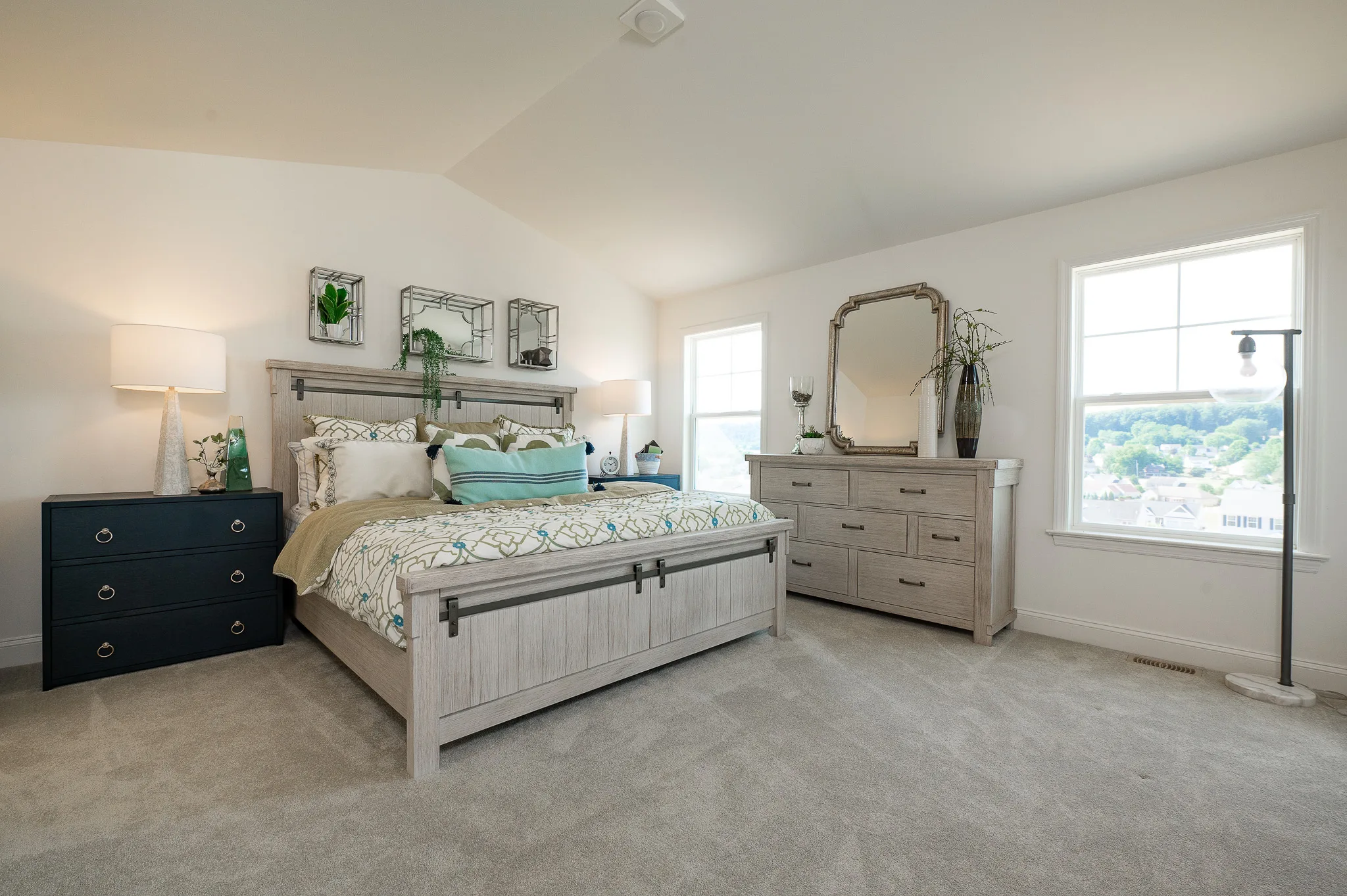 A bright, spacious bedroom at Eagle's View with a large bed, matching light wood dresser and nightstand, two table lamps, wall mirrors, and large windows letting in natural light. Neutral carpet and green accents complete the decor.