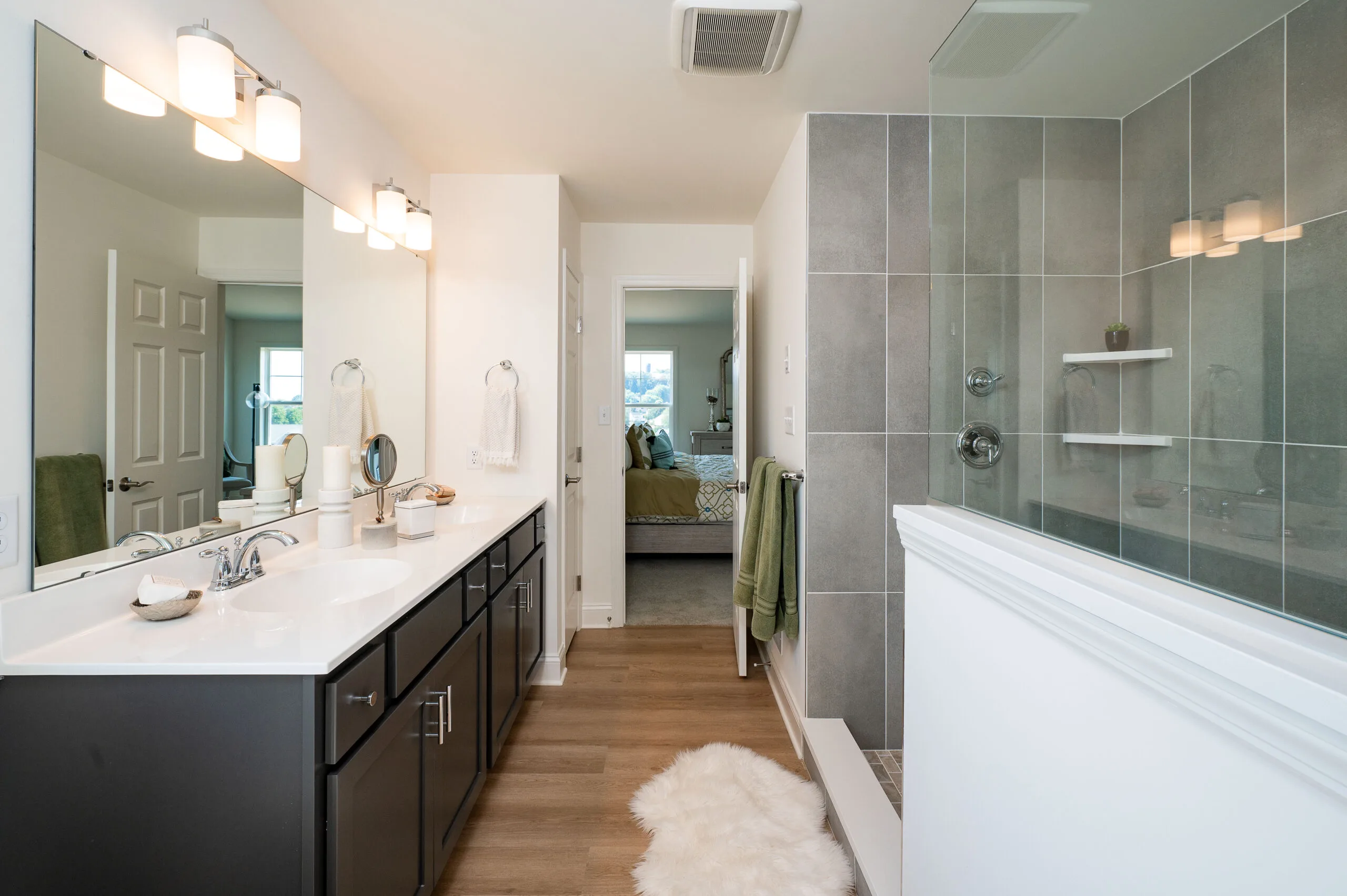 Modern bathroom with a double sink vanity, large mirror, and gray tile shower. There is a fluffy white rug on the wooden floor and a view into a bedroom through an open door. The space is bright and clean.