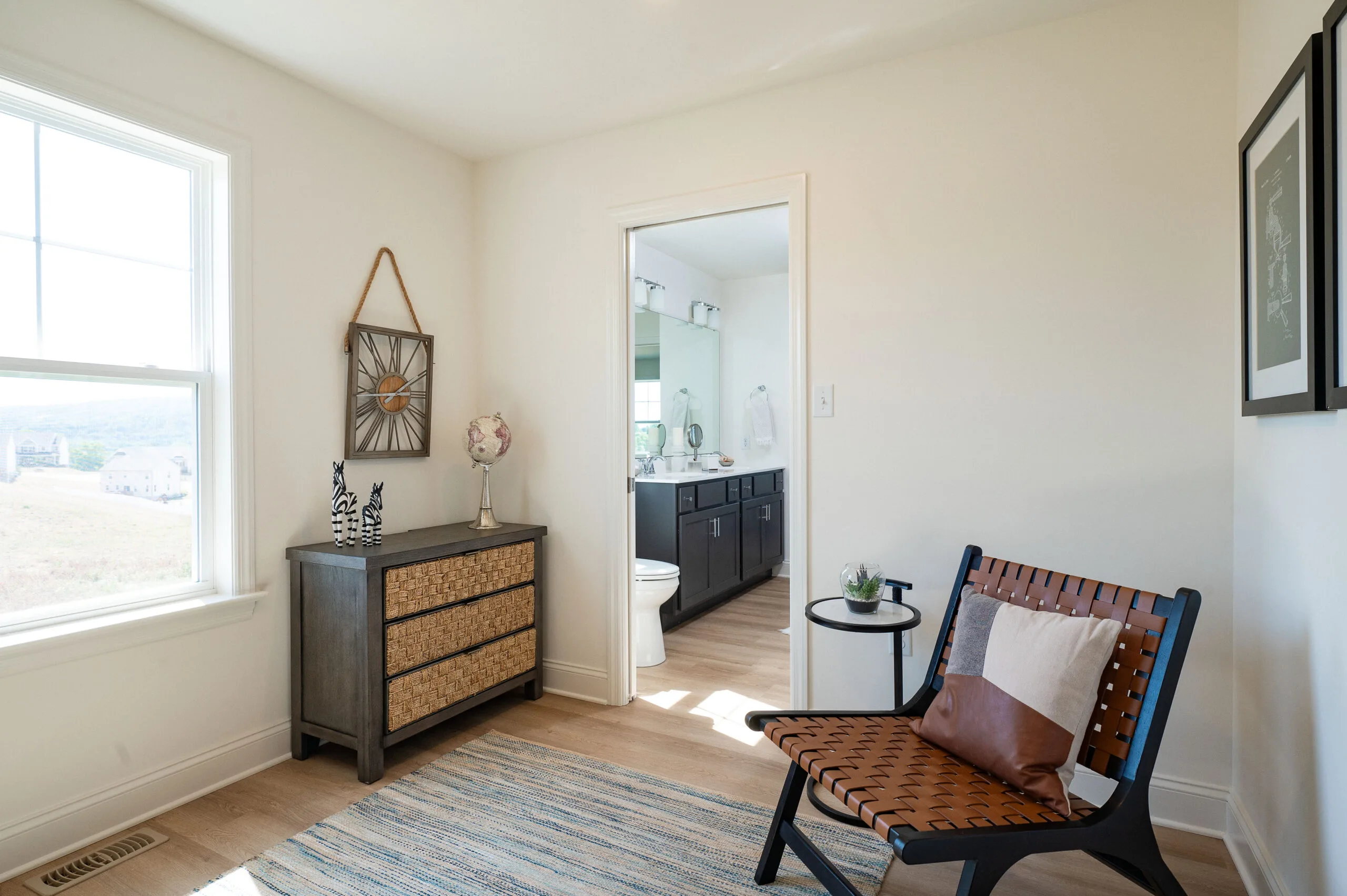A bright, modern bedroom with a woven chair, a striped rug, a small dresser with decorative items, and an open door leading to a bathroom with a double-sink vanity. Sunlight streams in through a large window.