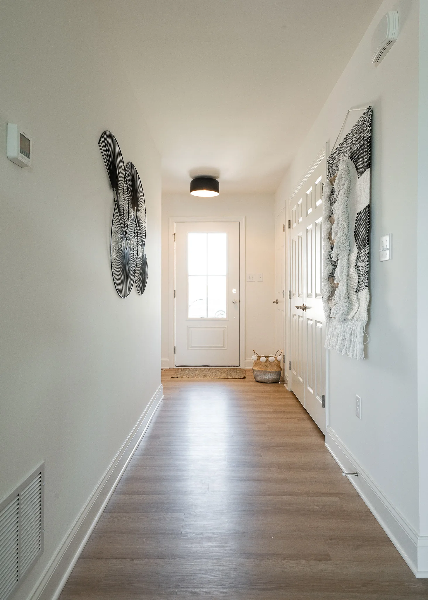 A bright hallway with light wood flooring, white walls, modern black wire wall art, a textured wall hanging, and a white door at the end. An American touch is added with a basket of blankets placed near the door for cozy comfort.