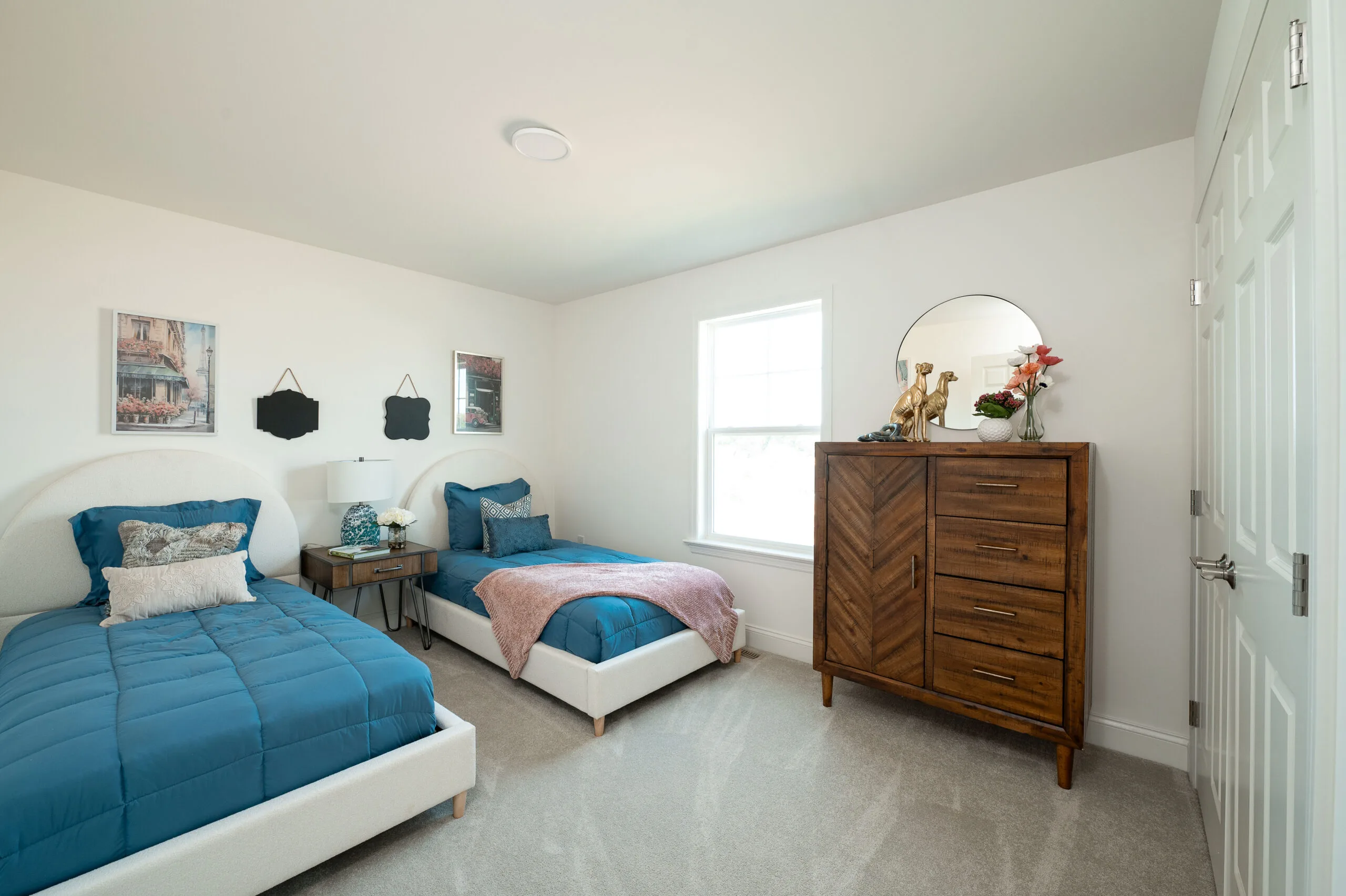 A bright bedroom with two twin beds with blue comforters, a nightstand between them, a window, wall art, and a wooden dresser with a round mirror and decorative items on top.