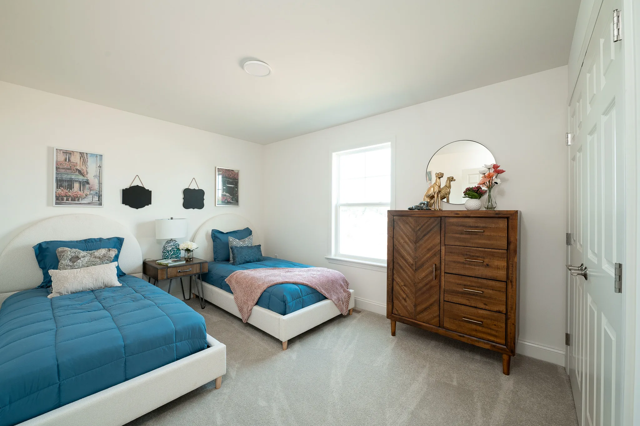 Two beds with blue bedding sit side by side in a bright Summergrove bedroom with white walls, a wooden dresser with a round mirror, and a window letting in natural light. Art and decor items accent the wall and dresser.