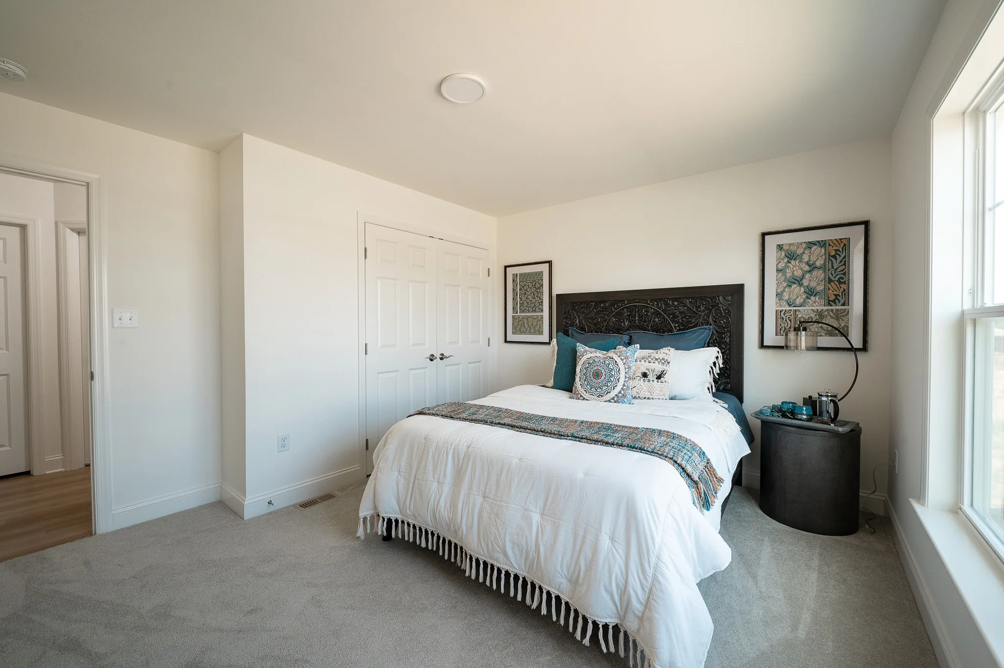A bright Summergrove bedroom with a neatly made bed featuring white linens and blue accents, two framed artworks above the headboard, a round black nightstand with decor, and a large window letting in natural light.