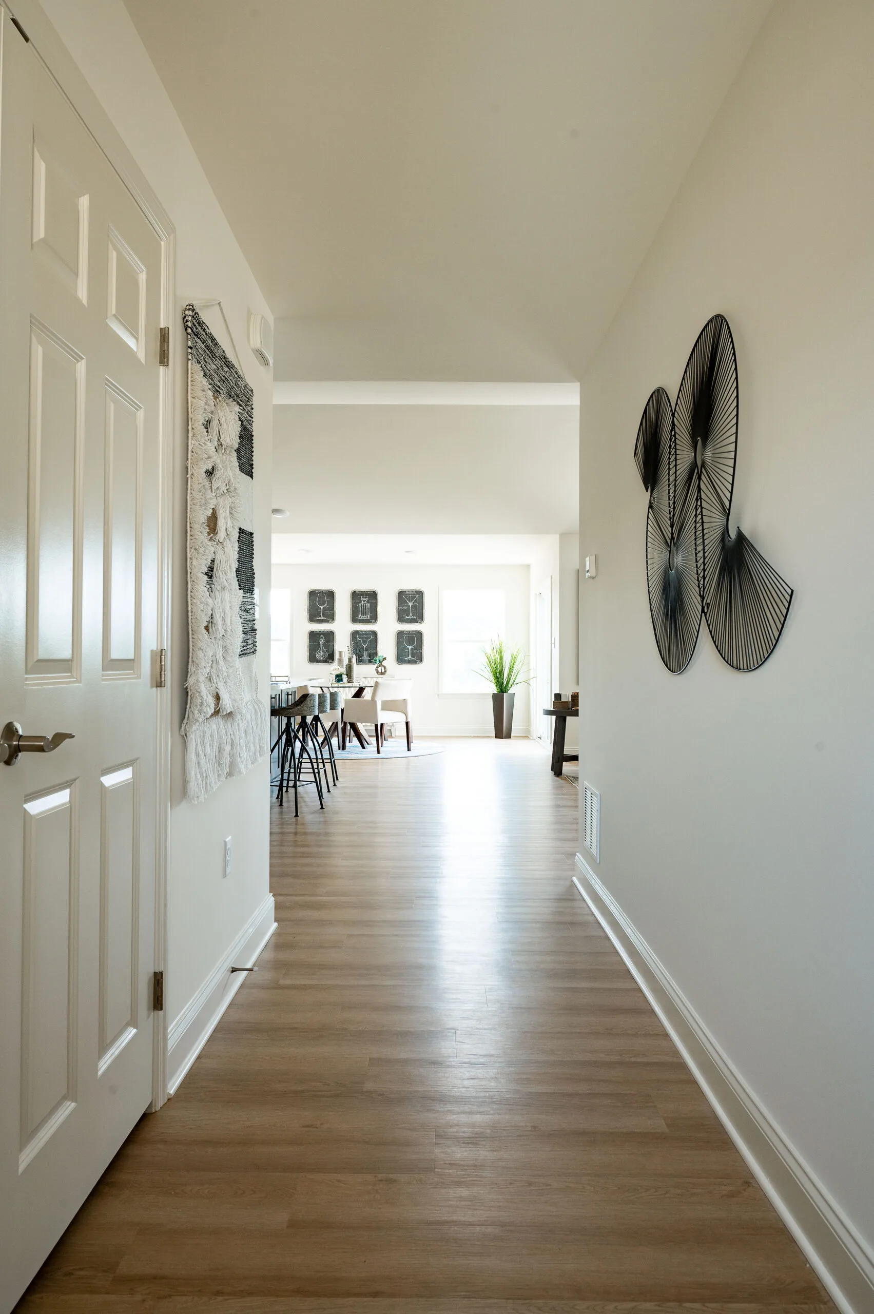 A hallway with light wood flooring leads to a bright, open dining area. Wall art decorates the hallway walls, and sunlight streams in from large windows near a potted plant in the dining space.