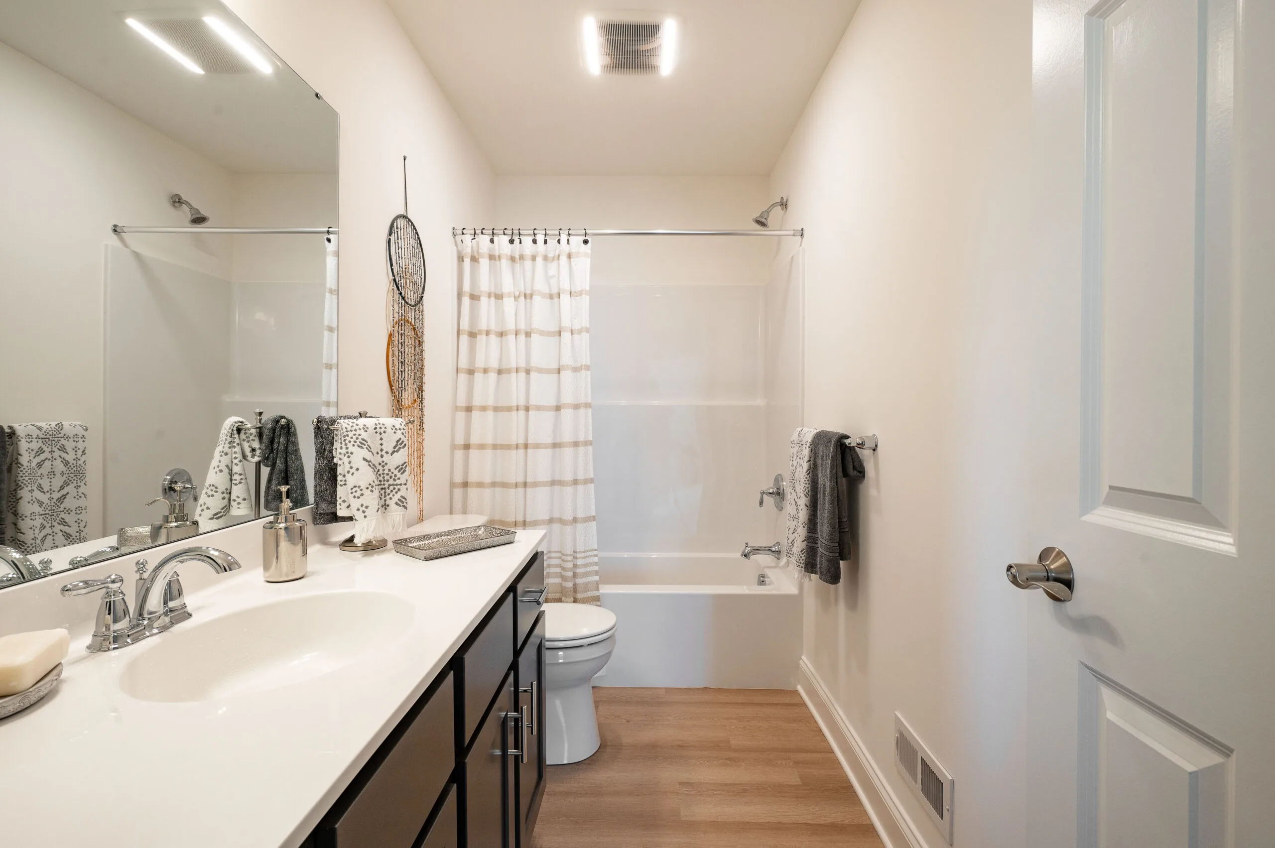 Modern bathroom with white walls, a large mirror above a sink with dark cabinets, silver fixtures, and a soap dispenser. A toilet and a bathtub with a striped shower curtain are in the background. Towels hang on the wall.