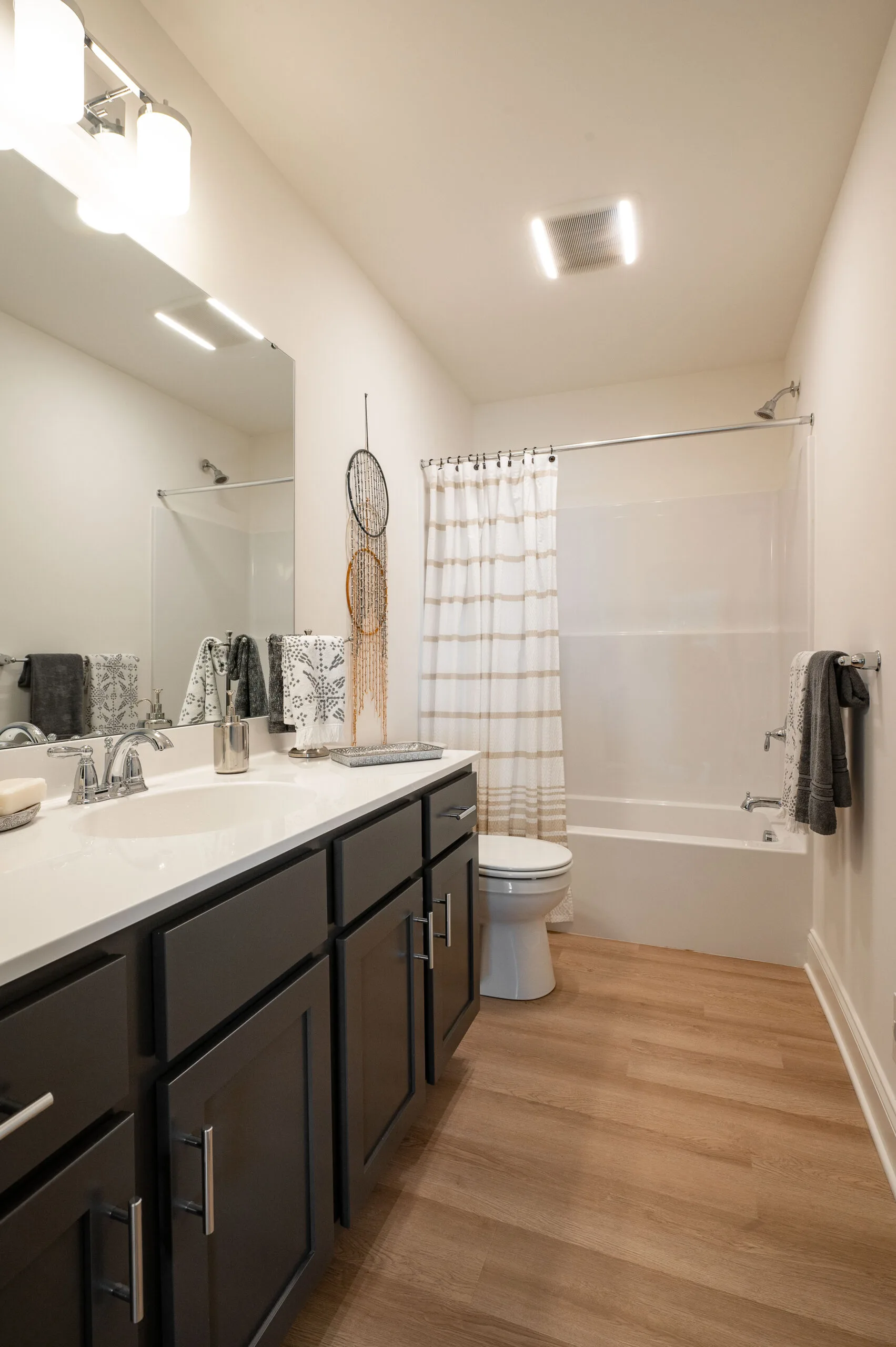 Modern bathroom with dark cabinets, white countertop, large mirror, and wood flooring. A toilet and bathtub with a white and beige striped shower curtain are at the far end. Towels and decor complete the look.