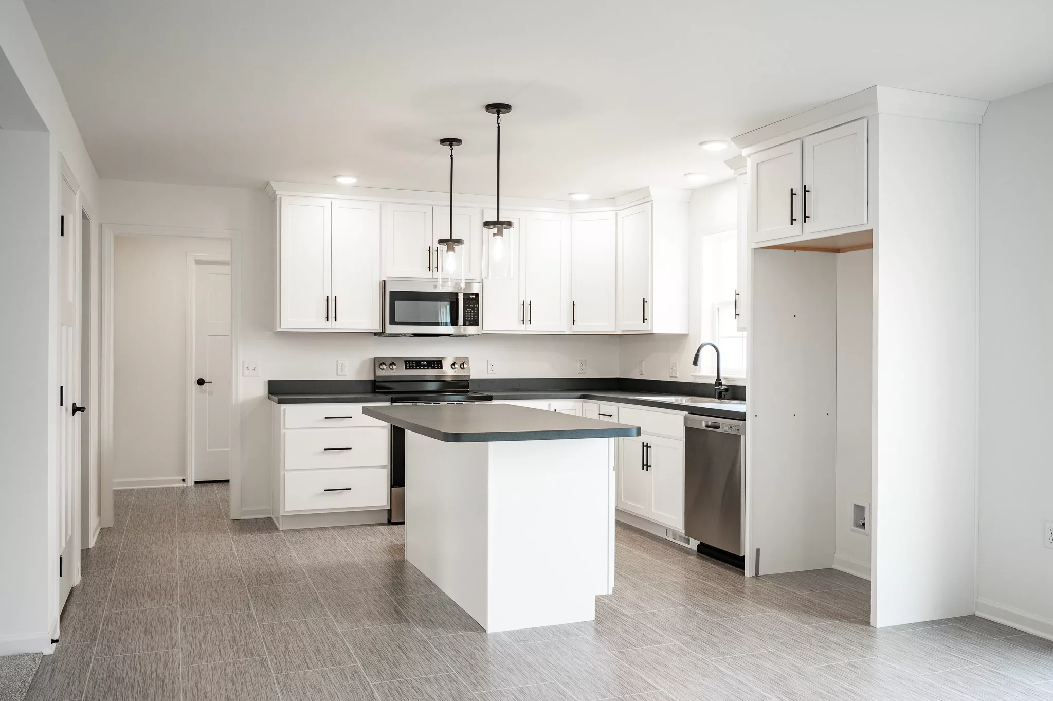 Modern kitchen with white cabinets, stainless steel appliances, a central island with a dark countertop, tile flooring, and pendant lights hanging above the island. The space has a clean, bright, and minimalist design.