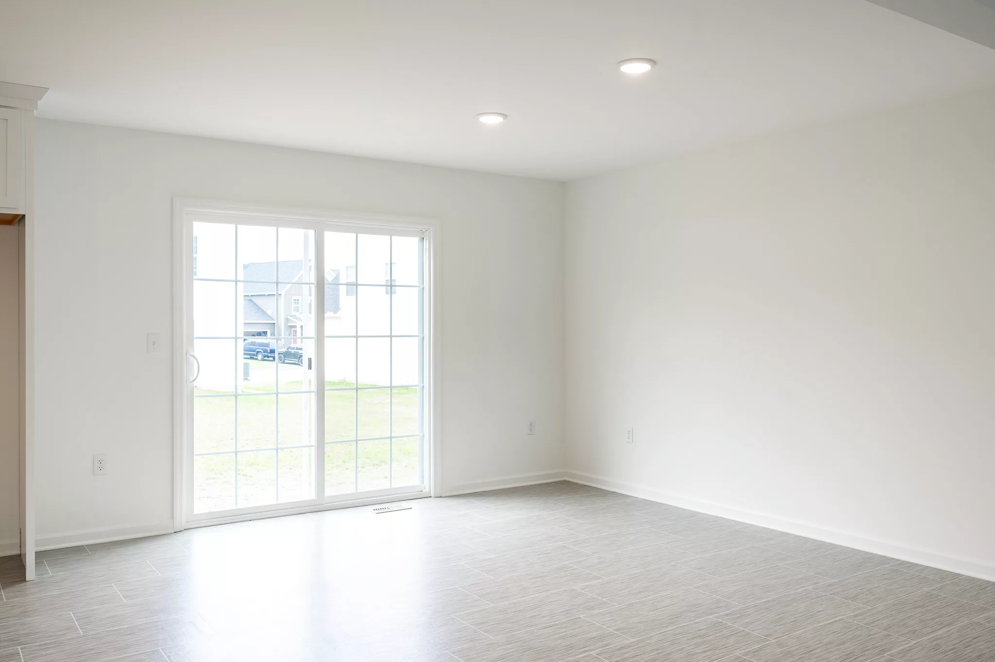 A bright, empty room with light-colored tile flooring, white walls, recessed ceiling lights, and large sliding glass doors leading to a grassy yard outside.