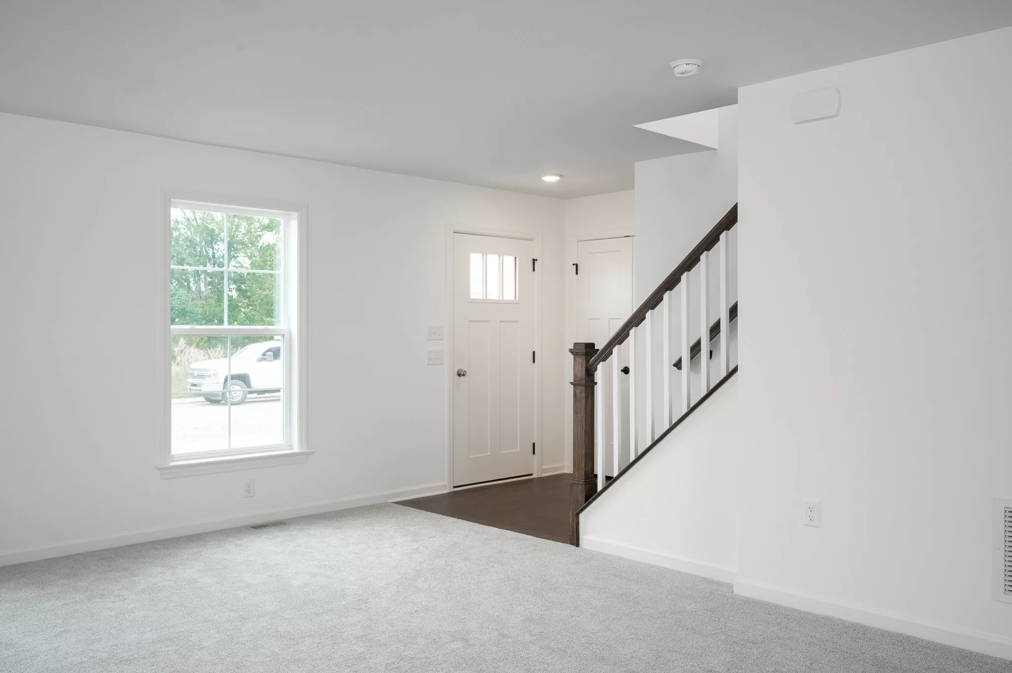 A bright, empty room with light gray carpet, a large window, a white front door, and a staircase with dark wooden handrails leading upstairs. Natural light fills the clean, minimal space.
