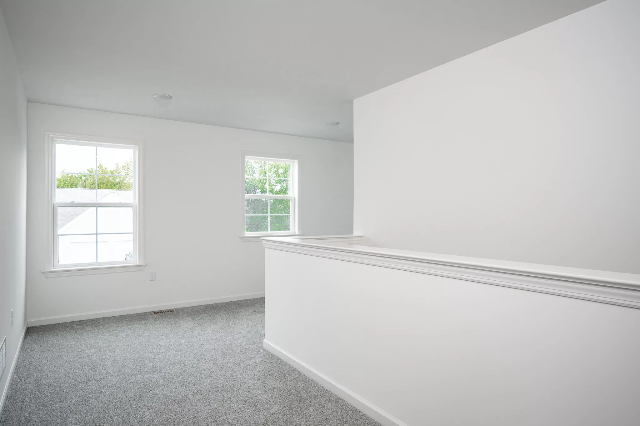 A bright, empty room with white walls, gray carpet, two windows letting in natural light, and a short white partition wall dividing the space.