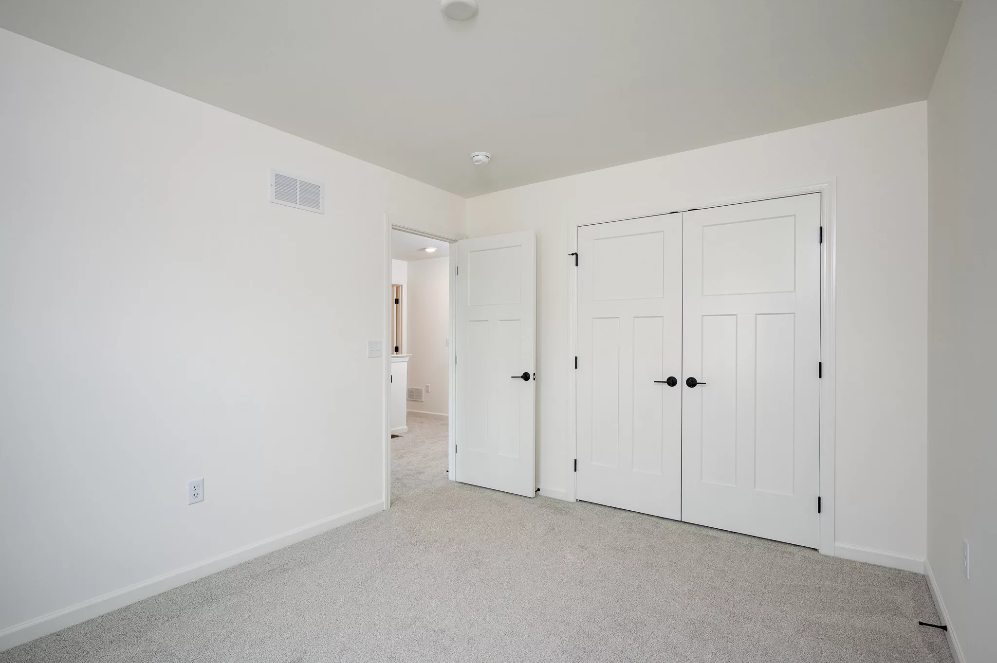 Empty bedroom with white walls, light carpet, and two closed white double doors with black handles. An open door leads to a hallway with similar light-colored decor. The room is well-lit with natural light.