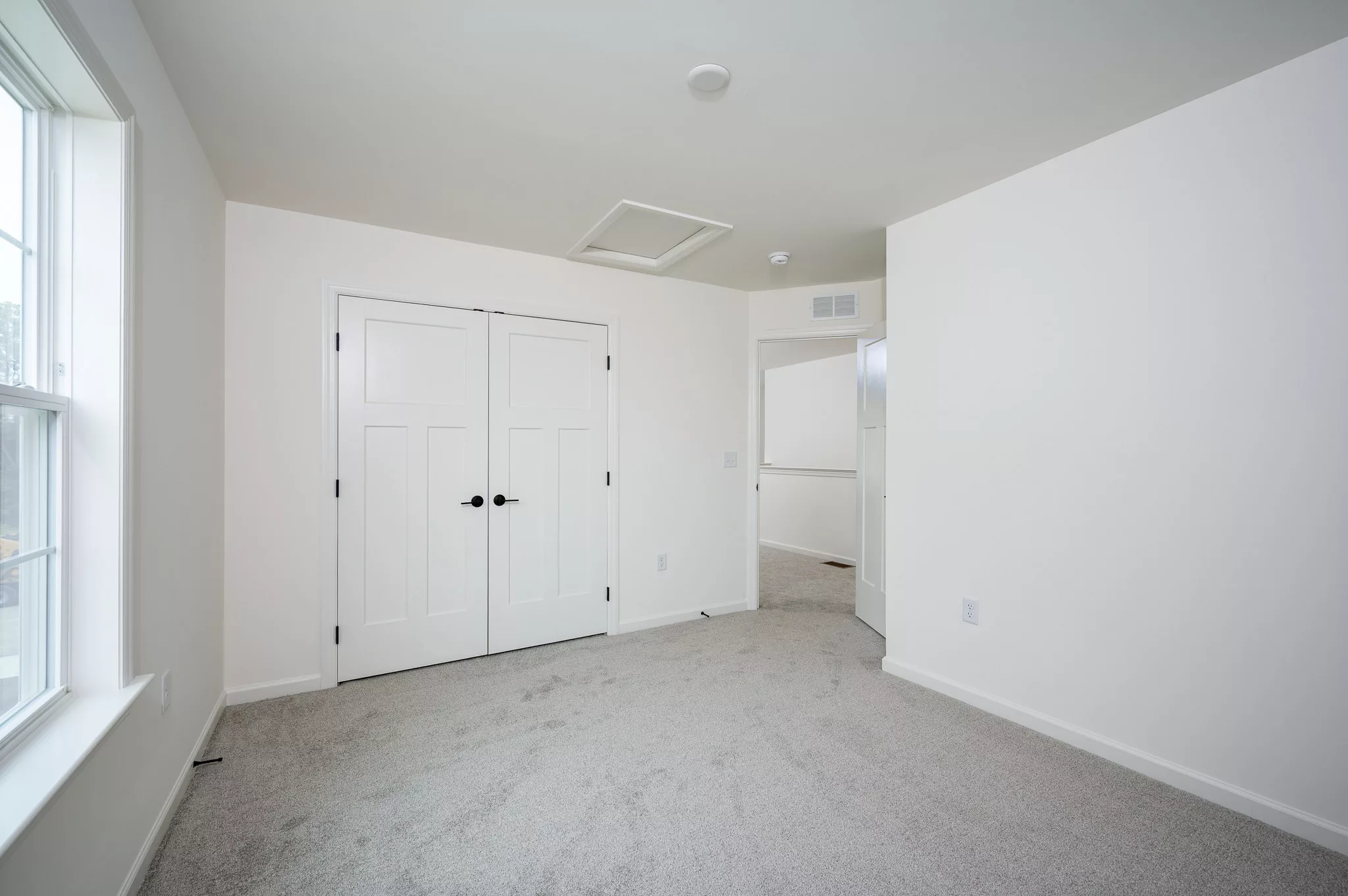 A bright, empty room with beige carpet, white walls, a large window on the left, double white closet doors with black handles, and an open doorway leading to a hallway.
