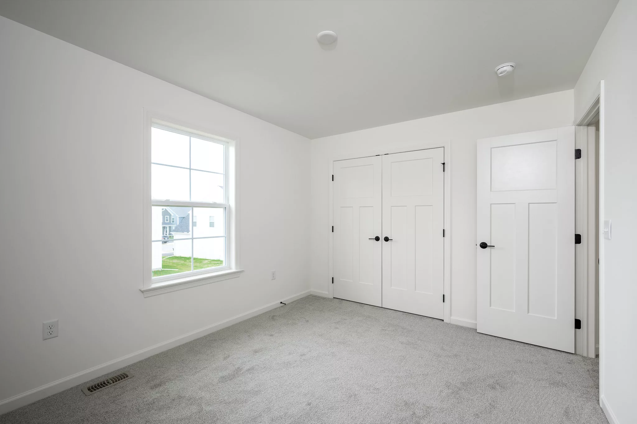 A bright, empty room with white walls, a gray carpet, a window letting in natural light, and two closed white double doors with black handles. One door on the right is slightly open.