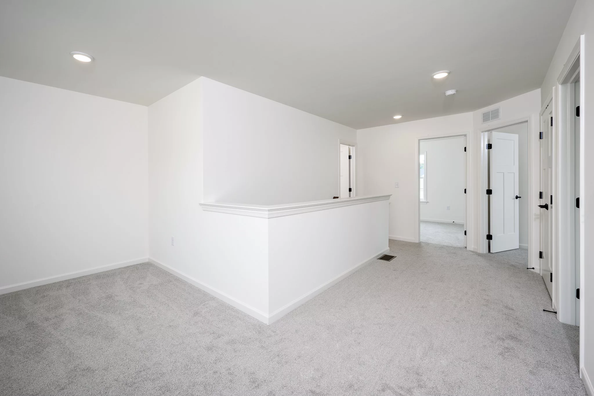 A bright, empty room with light gray carpet, white walls, recessed ceiling lights, and multiple doorways leading to other rooms. A half-wall and railing run along the center of the space.