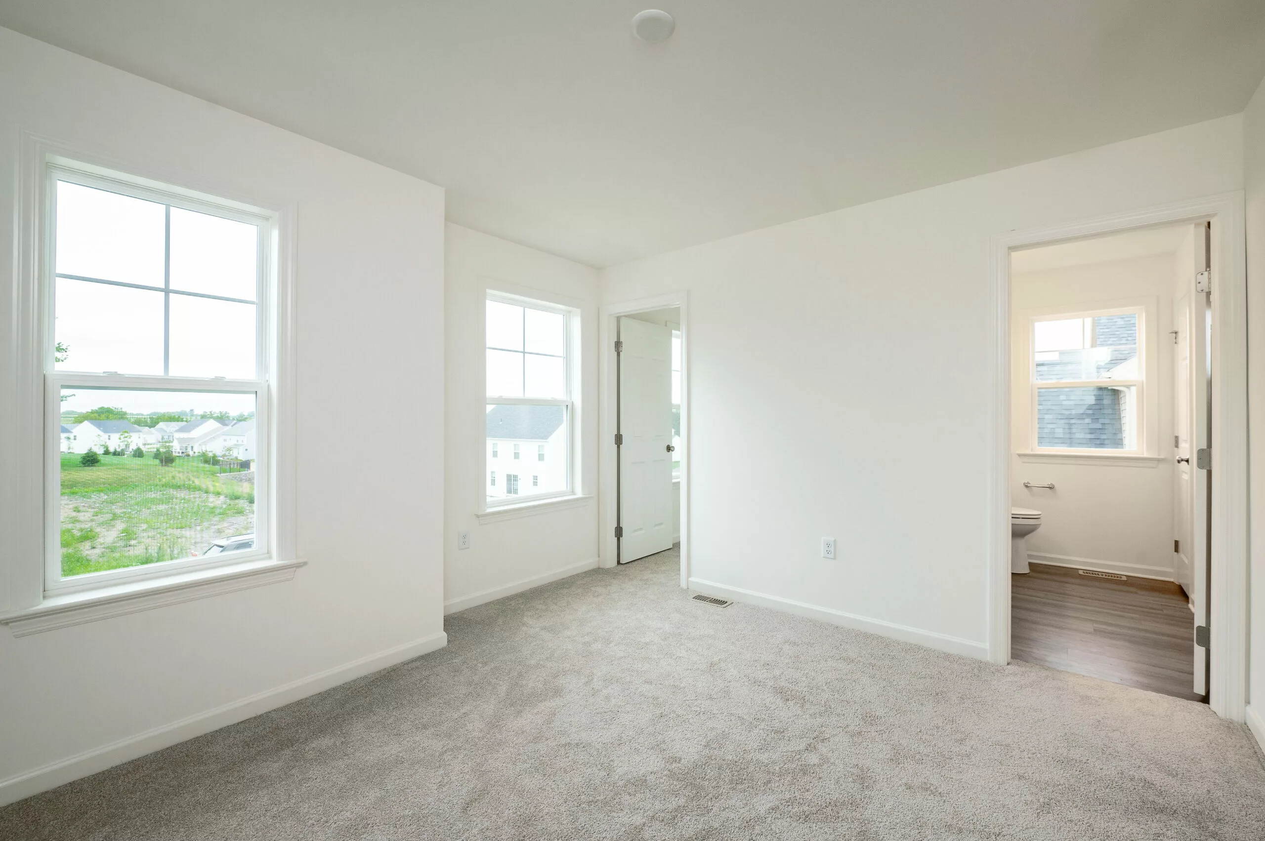 A bright, empty room with beige carpet, white walls, and two large windows. An open doorway leads to a small bathroom with a window and visible toilet. Natural light fills the space.