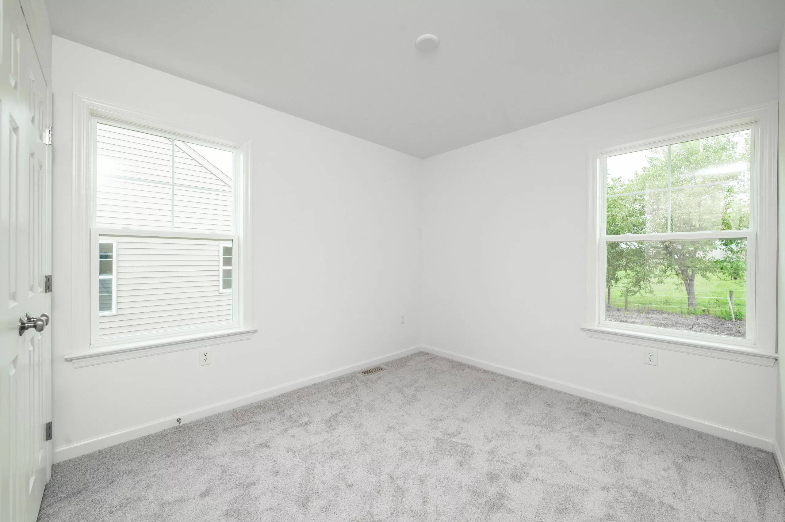 Empty, unfurnished room with white walls, light gray carpet, and two windows letting in natural light. One window shows the side of a house, the other shows green trees and grass outside.