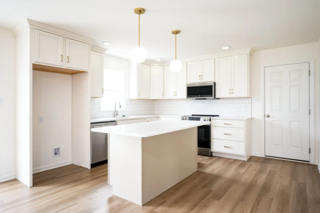 Modern kitchen with white cabinets, a central island, wood flooring, stainless steel appliances, and two round pendant lights hanging from the ceiling. There is an empty space for a refrigerator on the left.