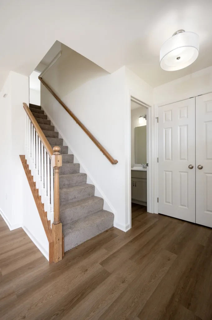 A bright hallway with wooden floors, carpeted stairs with a wooden handrail, double white closet doors, and an open door revealing a bathroom sink and mirror. A round ceiling light hangs above.