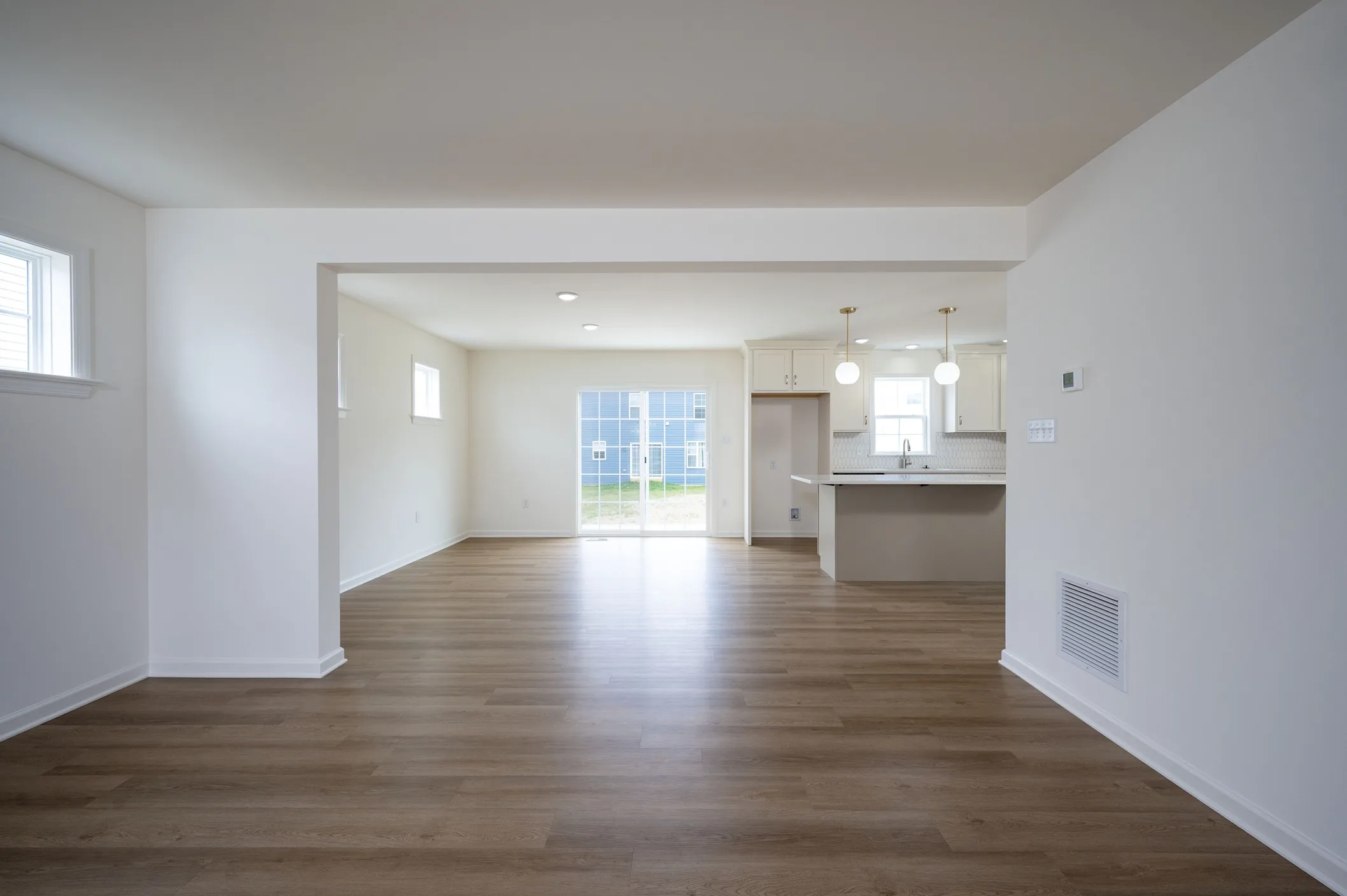 Spacious, empty living area with light wood floors, white walls, and large windows. In the background, a kitchen with pendant lights and sliding glass doors leading outside are visible.