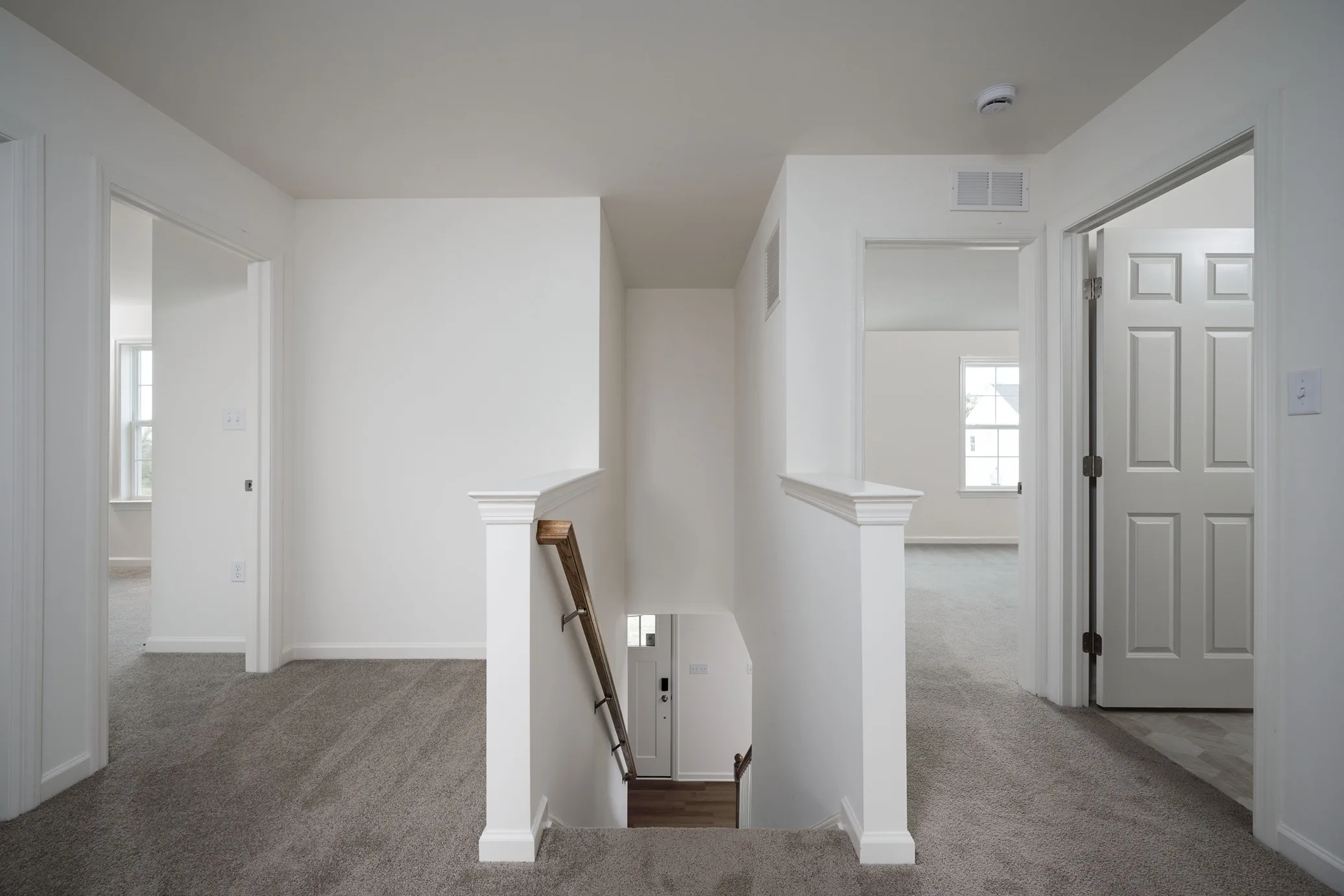 A carpeted upstairs hallway with a staircase and three doorways leading to bright, empty rooms with white walls and large windows.