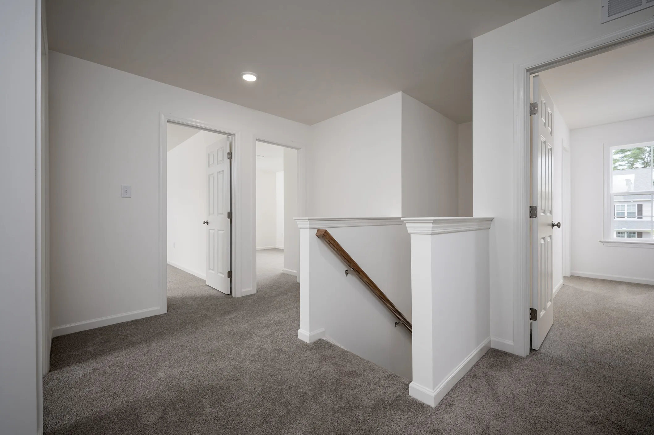 A hallway with gray carpet and white walls, featuring three open doors leading to different rooms and a staircase with a wooden handrail descending to a lower level. Natural light enters through a window in one of the rooms.