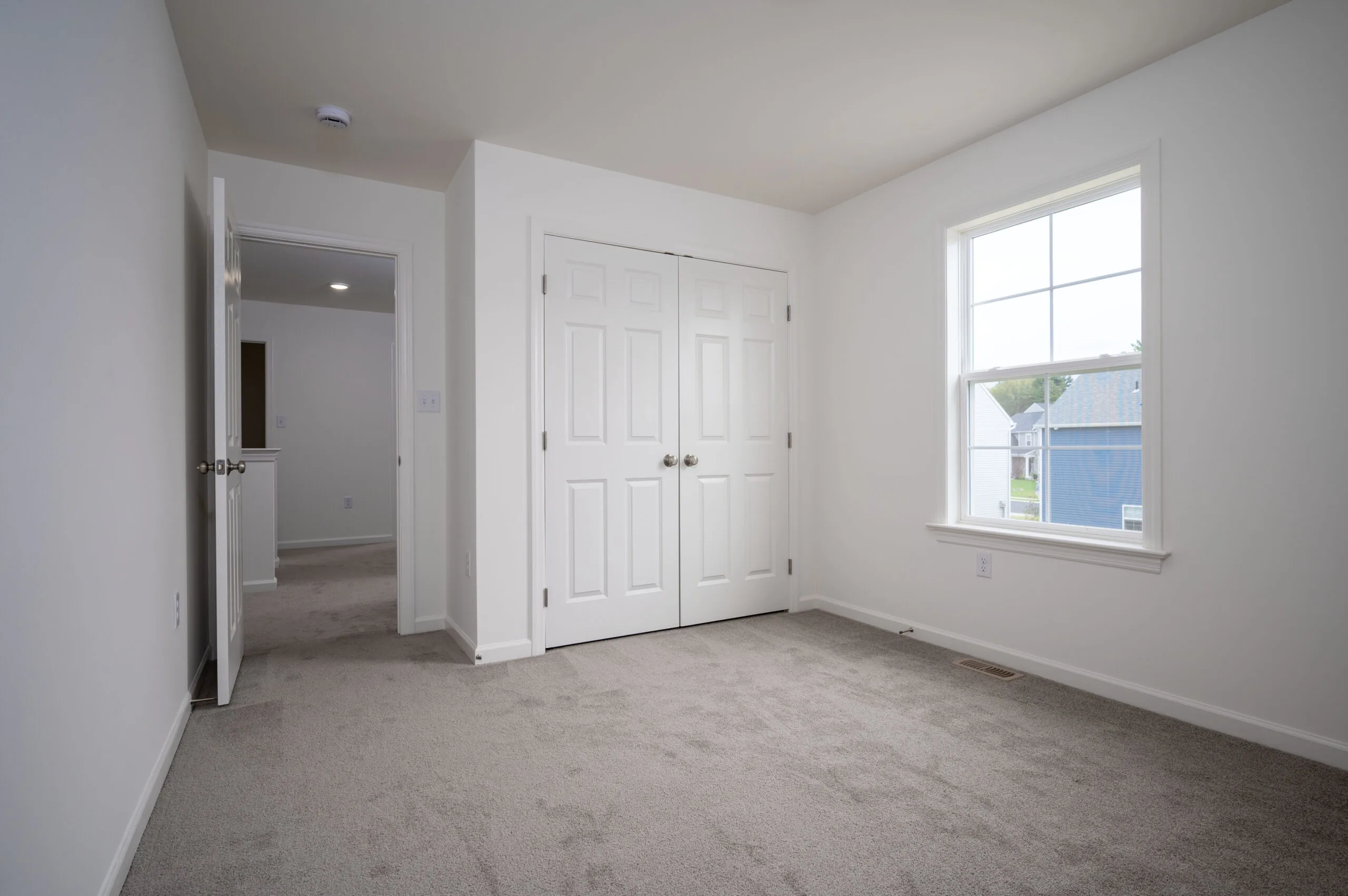 A small, empty bedroom with beige carpet, white walls, a closed double closet with white doors, an open door leading to a hallway, and a window letting in natural light.