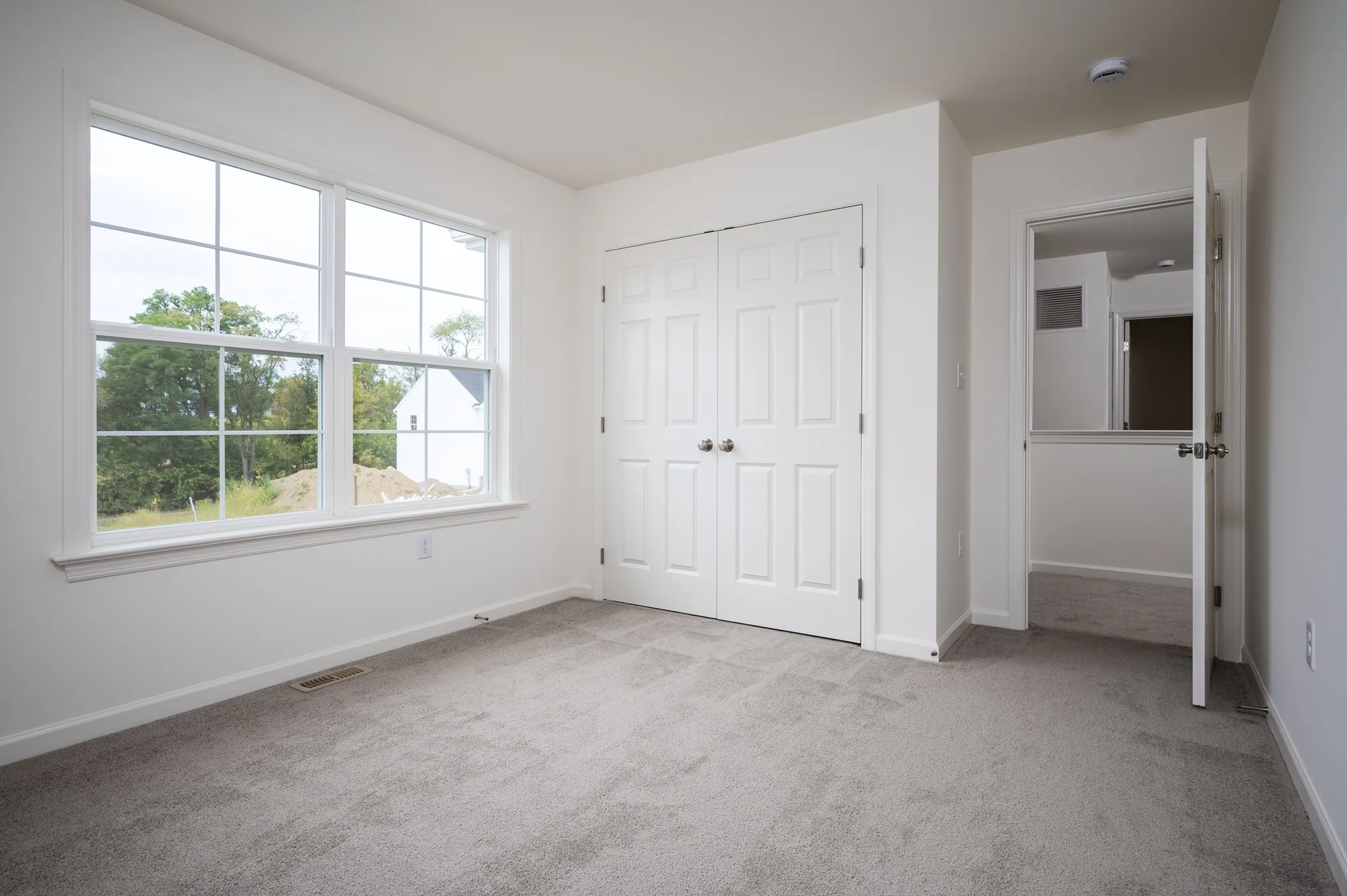 Empty room with light gray carpet, white walls, a large window, double-door closet, and an open door leading to a hallway. Natural light brightens the space.