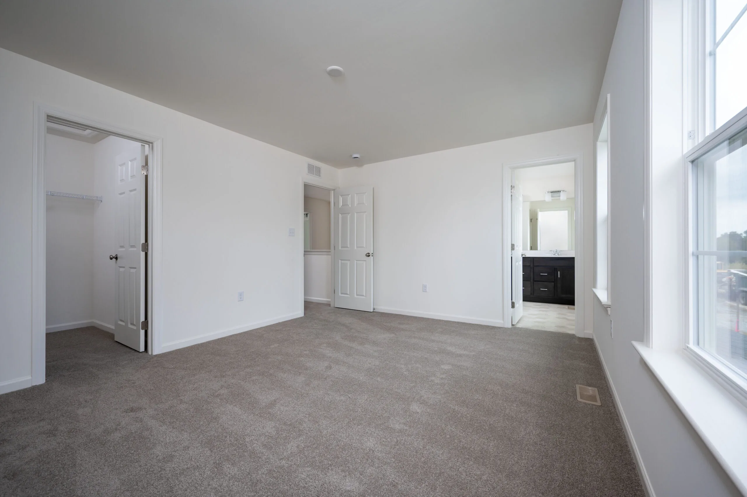 A spacious, empty bedroom with beige carpet, white walls, an open closet door on the left, a door to a hallway in the center, an open bathroom door on the right, and large windows letting in natural light.