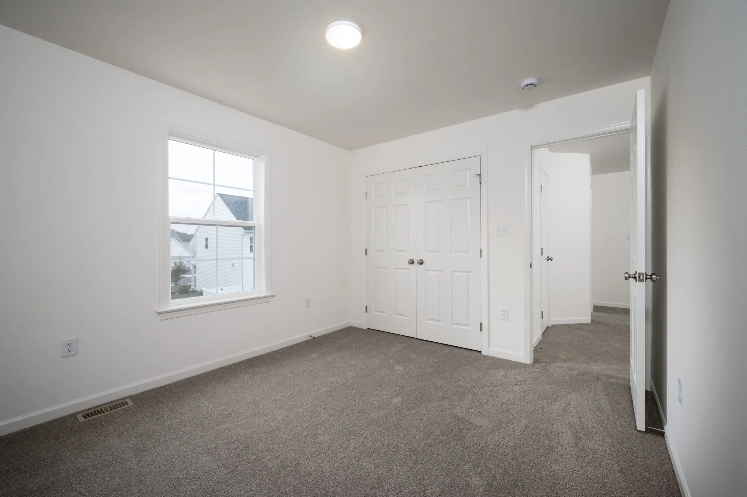 Empty bedroom with beige carpet, white walls, a window, a double-door closet, and an open door leading to a hallway. The room is well-lit with a ceiling light and natural daylight from the window.