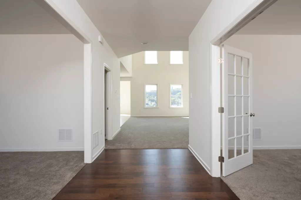 Bright, empty interior of a modern Lawrenceville home with white walls, hardwood floors leading to carpeted rooms, large windows, and glass French doors. Natural light fills the space, highlighting a clean, open layout.