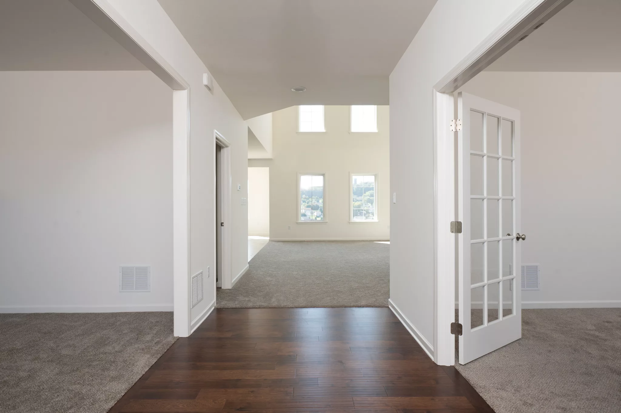 Bright, empty interior of a modern Lawrenceville home with white walls, hardwood floors leading to carpeted rooms, large windows, and glass French doors. Natural light fills the space, highlighting a clean, open layout.